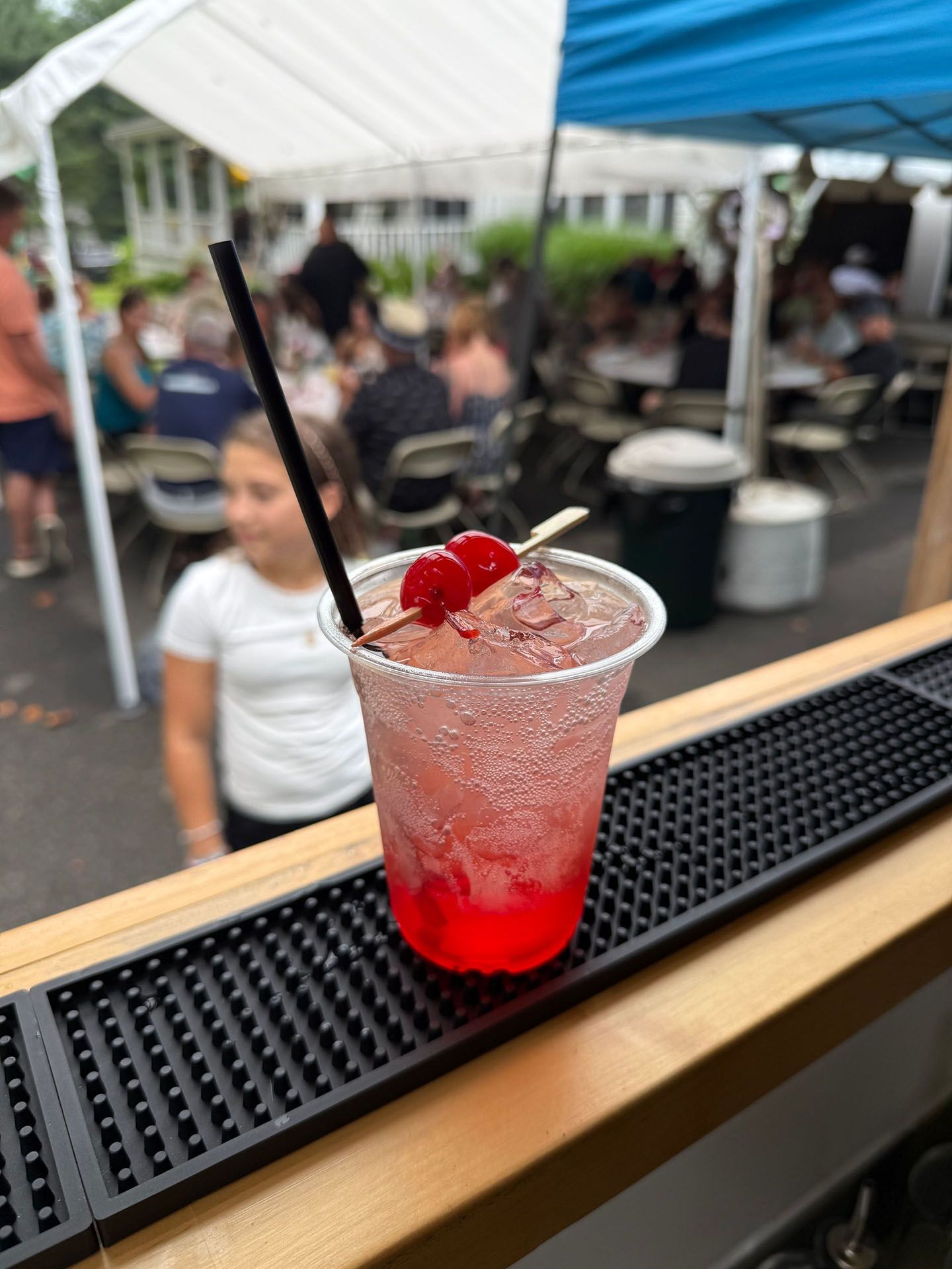 A red cocktail in a clear plastic cup, garnished with a cherry, on a bar; people and tents in the background.
