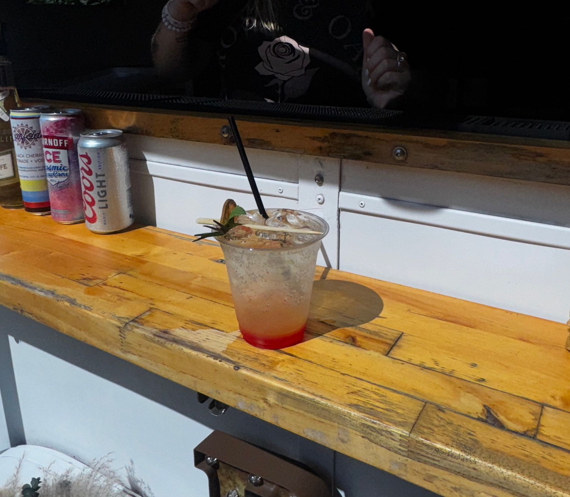 A cocktail in a clear plastic cup sits on a wooden bar, next to beer cans.
