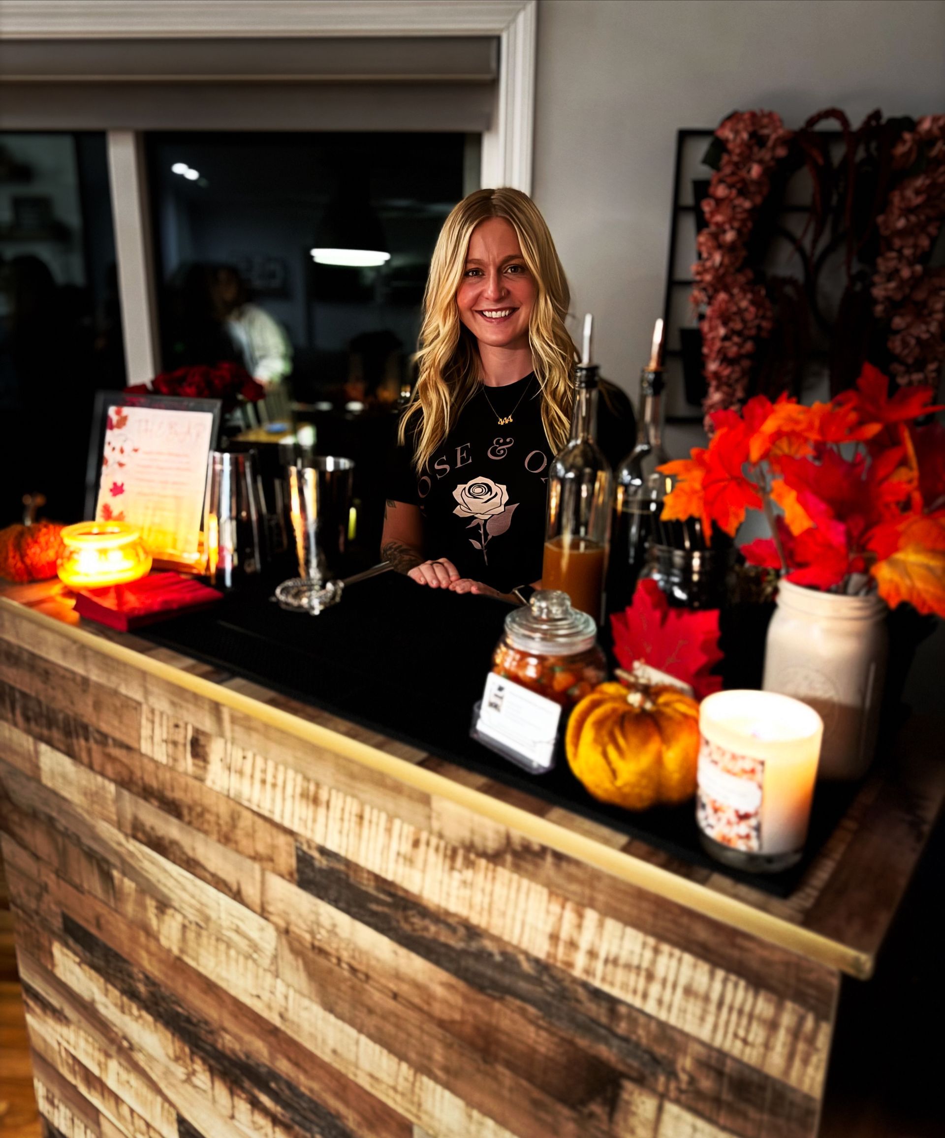 Woman behind a wooden bar, smiling. Fall decor includes pumpkins, candles, and autumn leaves.