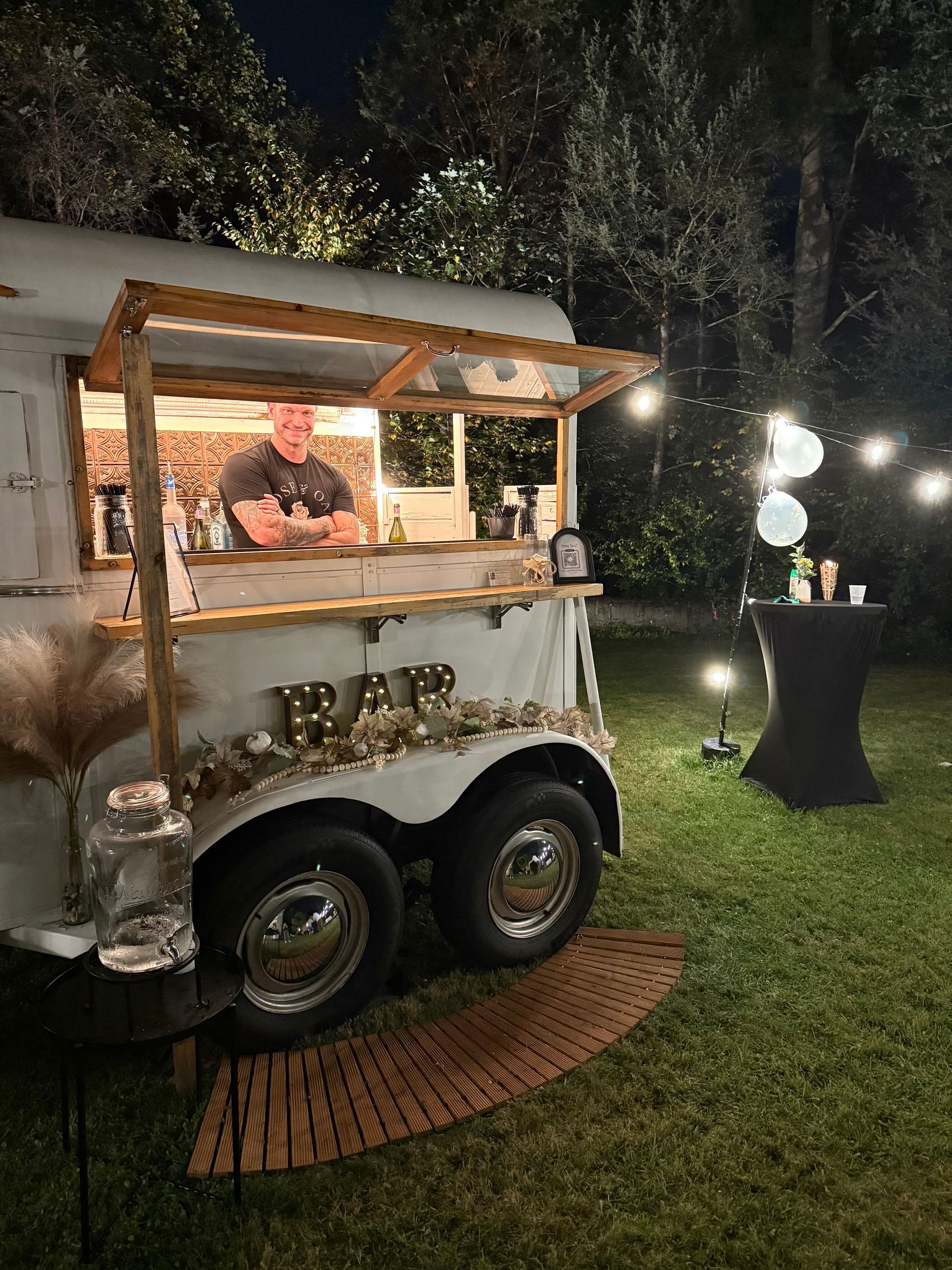 A mobile bar, set up outdoors at night. A bartender stands at the entrance, surrounded by string lights, and a small table with drinks nearby.