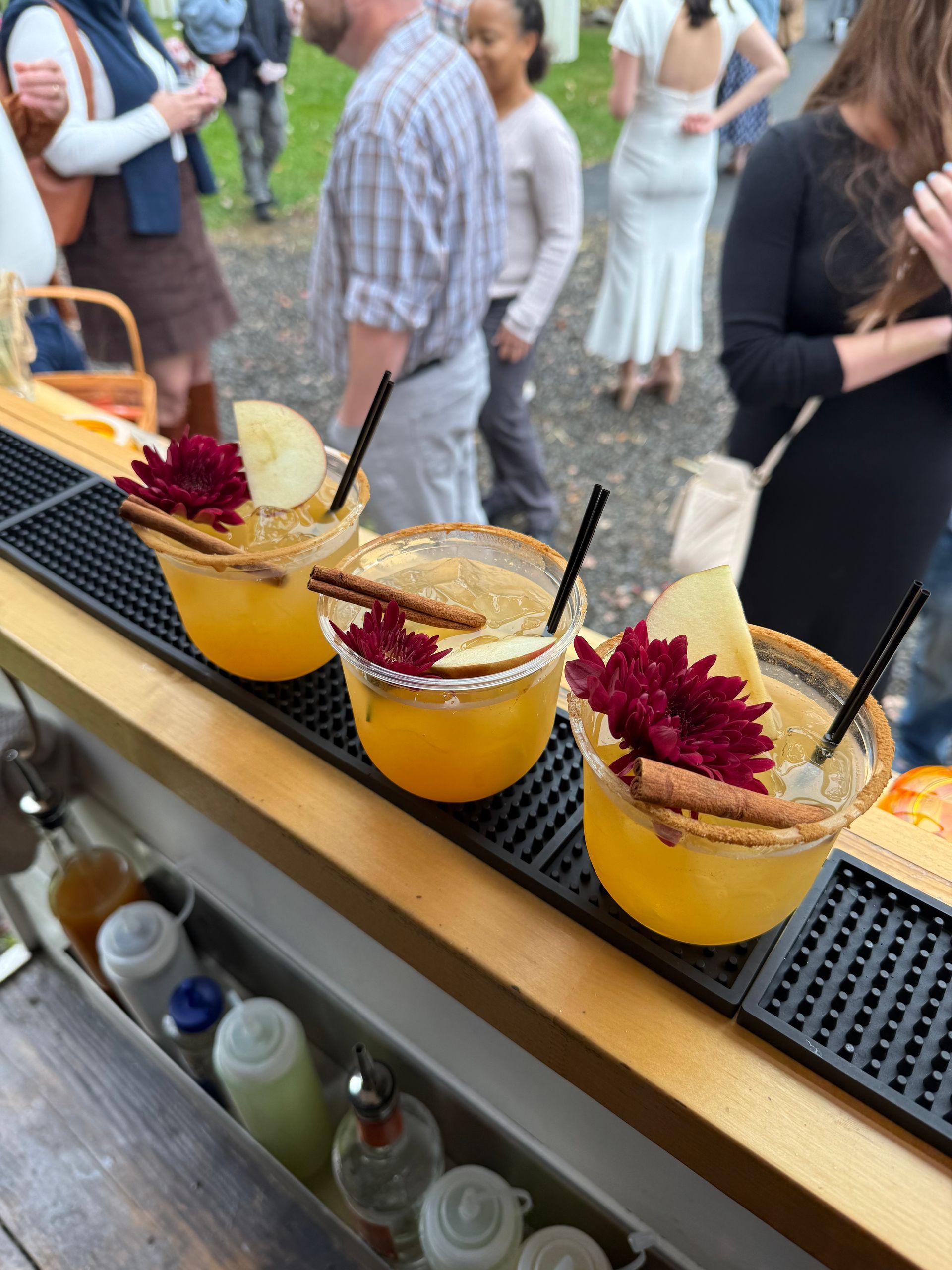 Three cocktails with apple slices, cinnamon sticks, and flowers on a bar, with people in the background.