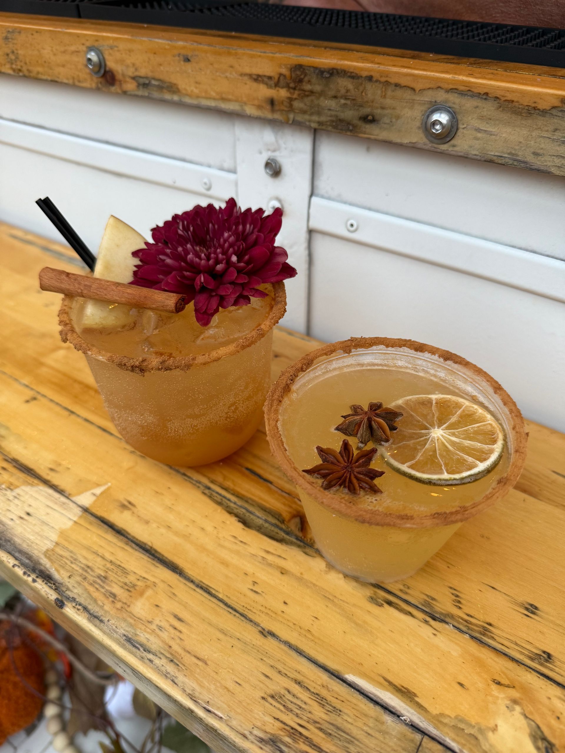 Two cocktails in cinnamon sugar-rimmed glasses, garnished with a flower, star anise, apple slice, and lime, on a wooden surface.