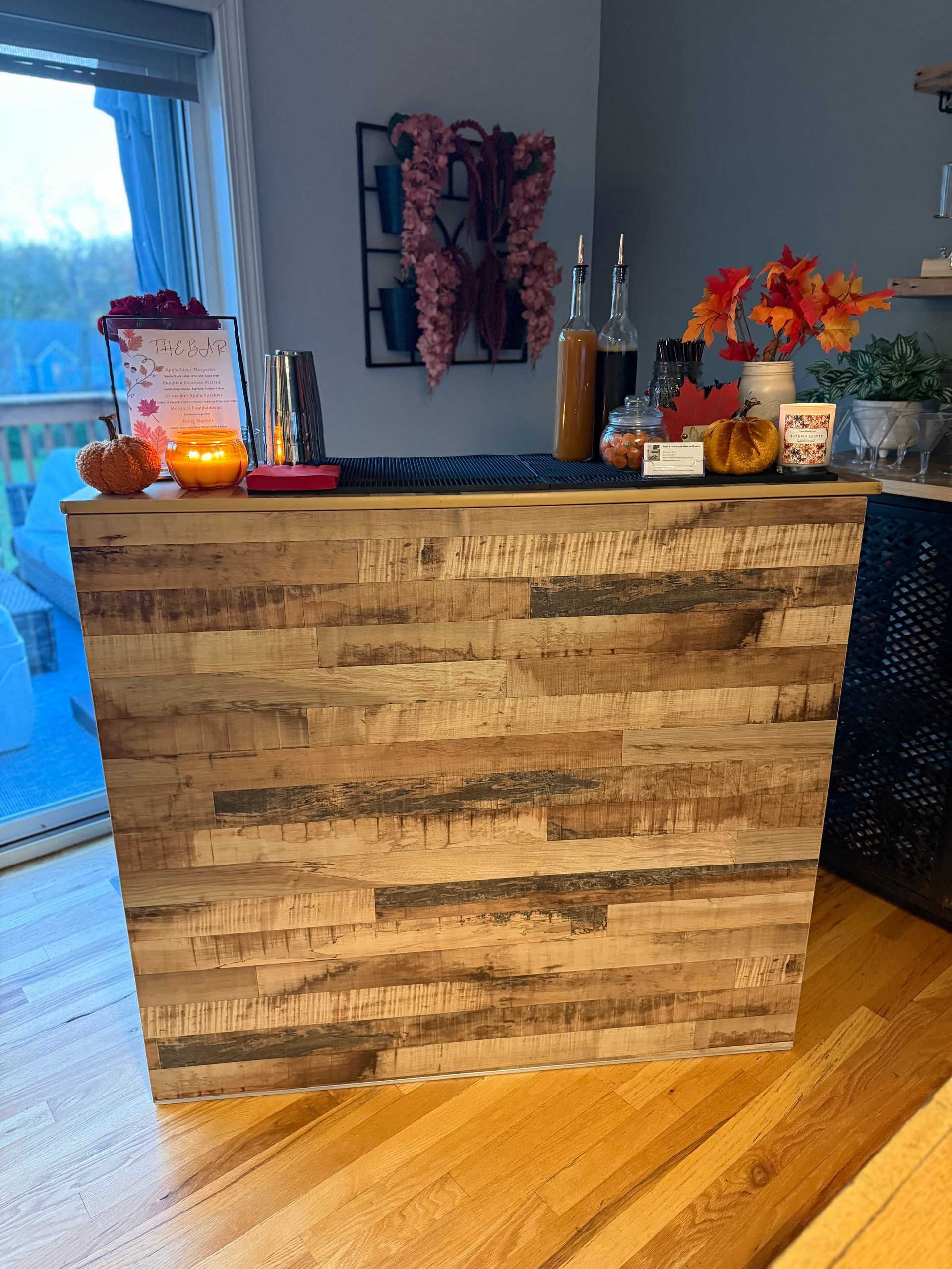 A wooden bar decorated with fall elements, featuring a lit candle, pumpkins, and autumn leaves.