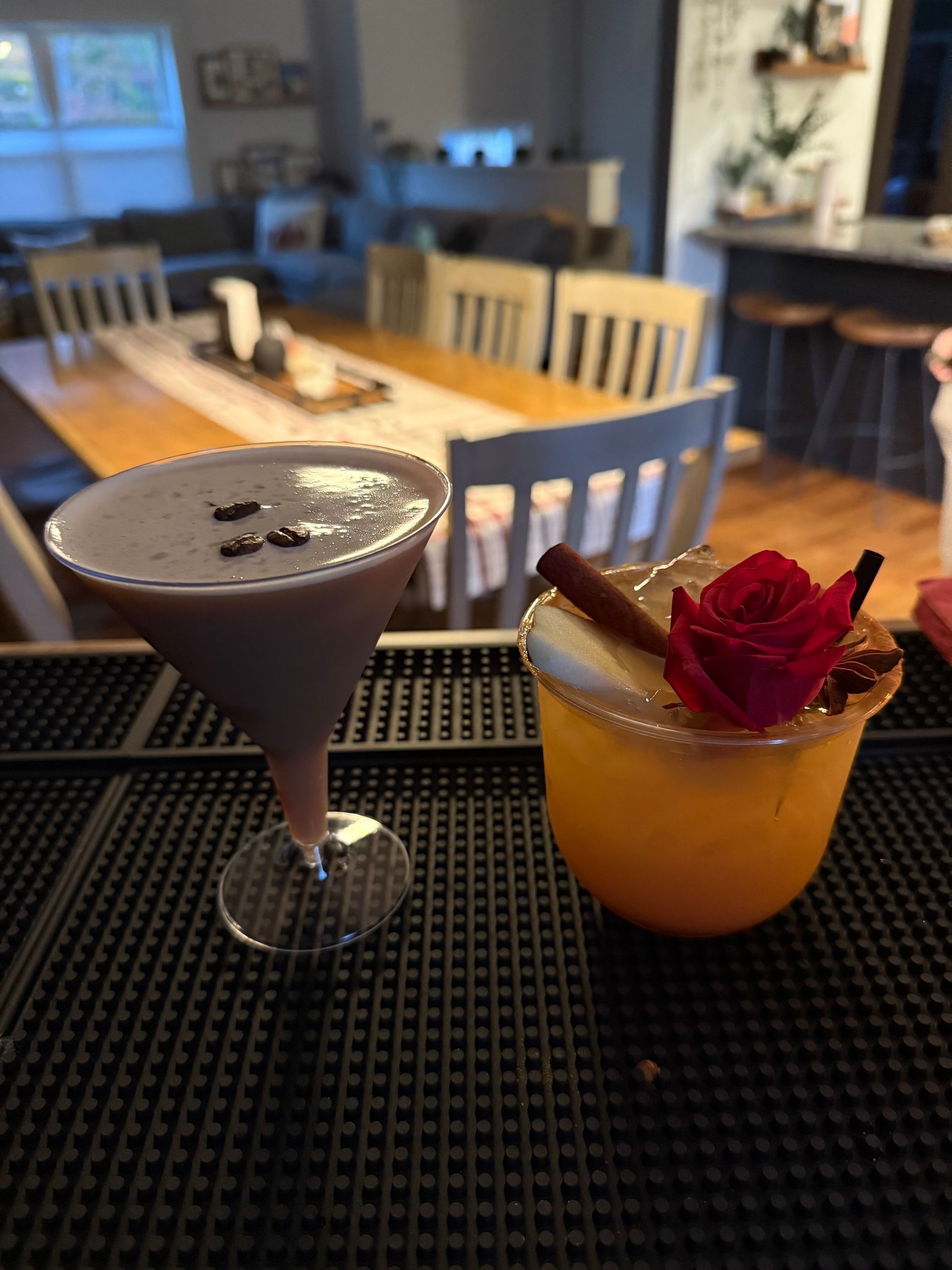 Two cocktails on a black bar mat: a chocolate martini with coffee beans and a yellow drink with a cinnamon stick and rose garnish.