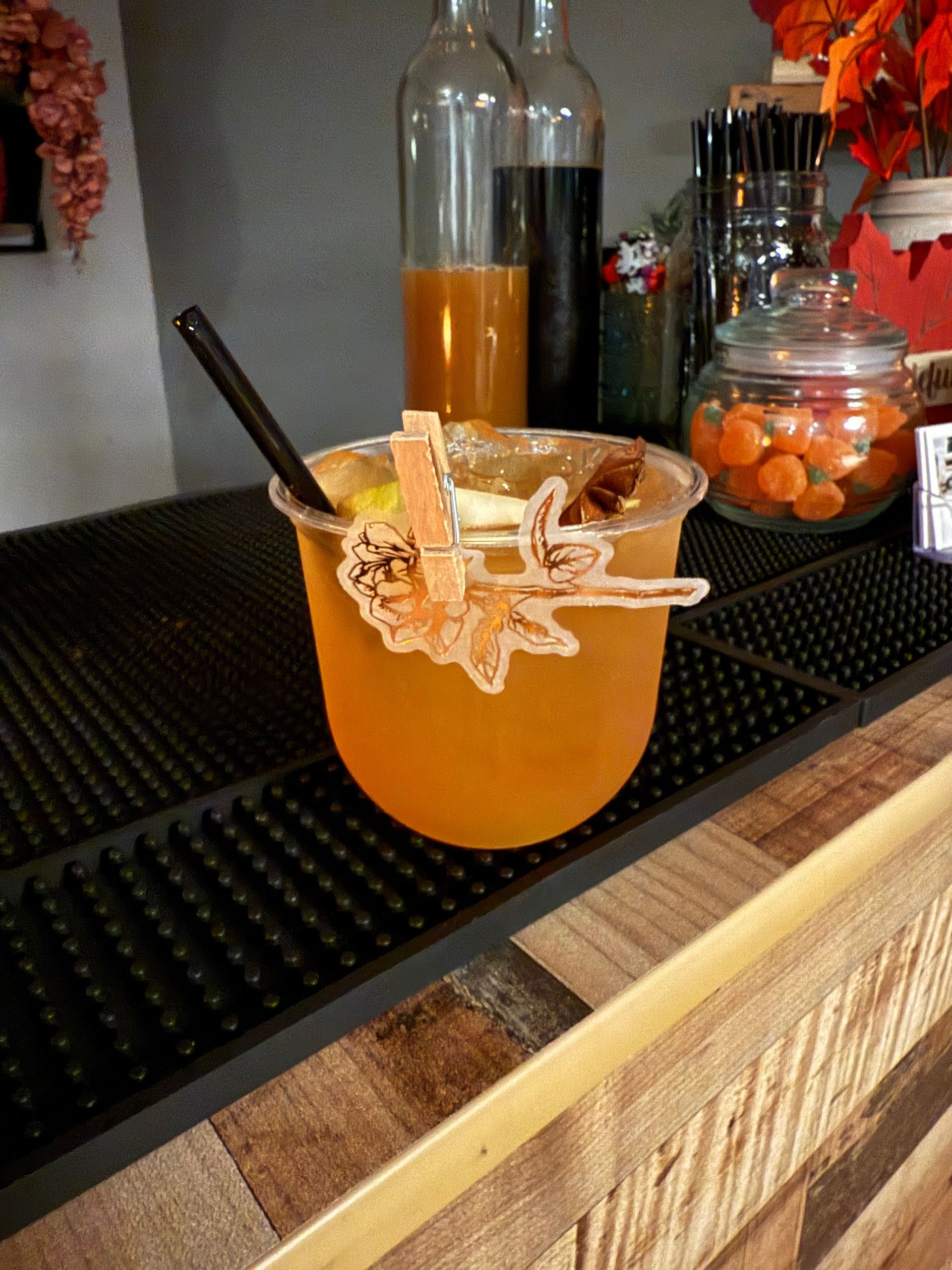 Cocktail in a clear glass with a straw and cinnamon stick garnish on a bar with bottles and a jar of carrots in the background.