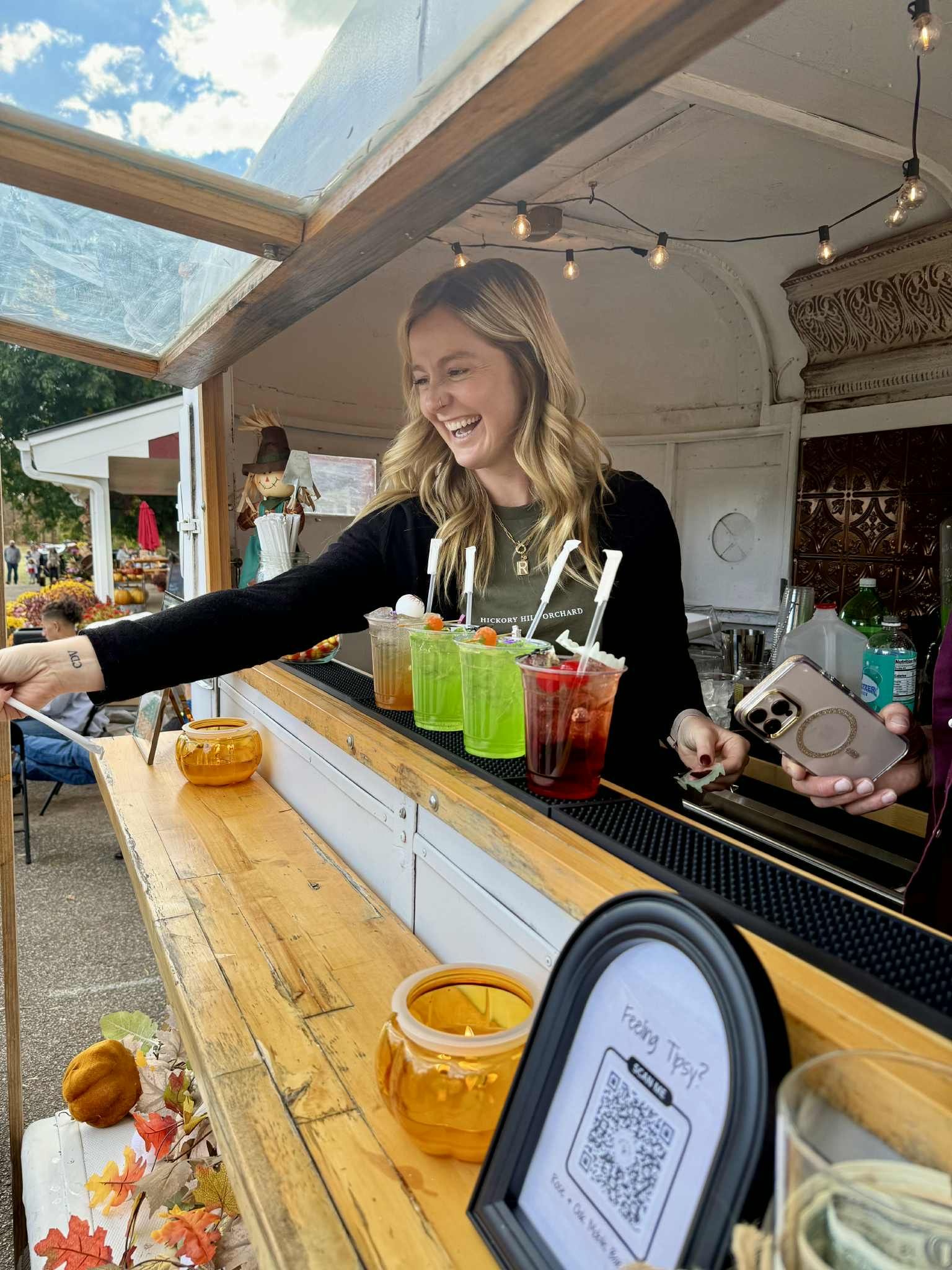 A woman laughing behind a bar, serving colorful drinks outdoors. Fall decorations and a QR code are visible.