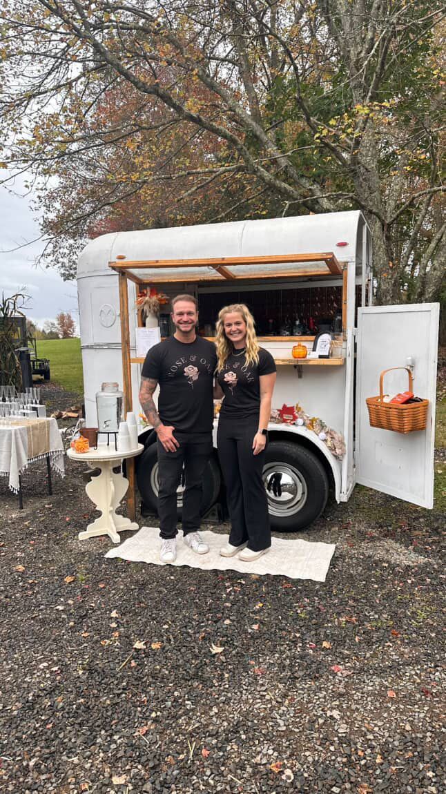 Two people stand in front of a white food trailer with a rustic wooden awning.