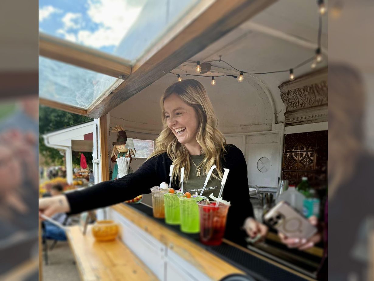 A woman smiling behind a bar, serving colorful drinks outside.