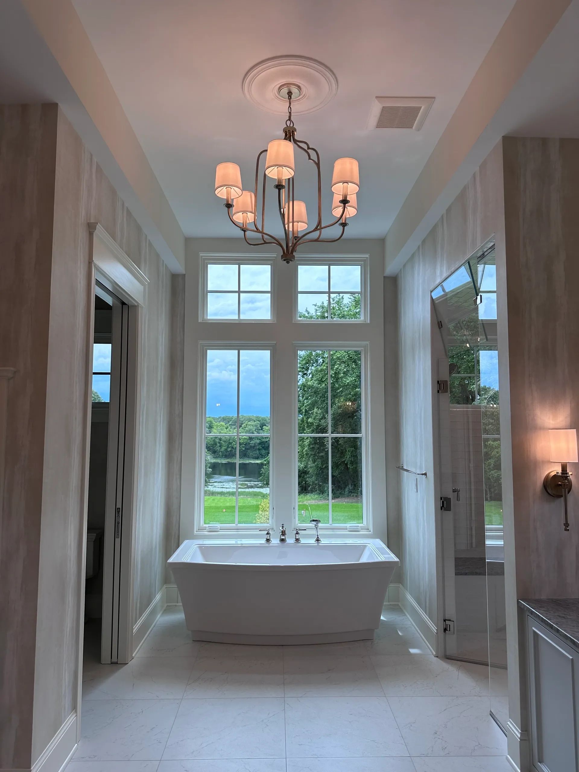 Elegant bathroom with freestanding tub under a large window, chandelier, and neutral-toned walls.