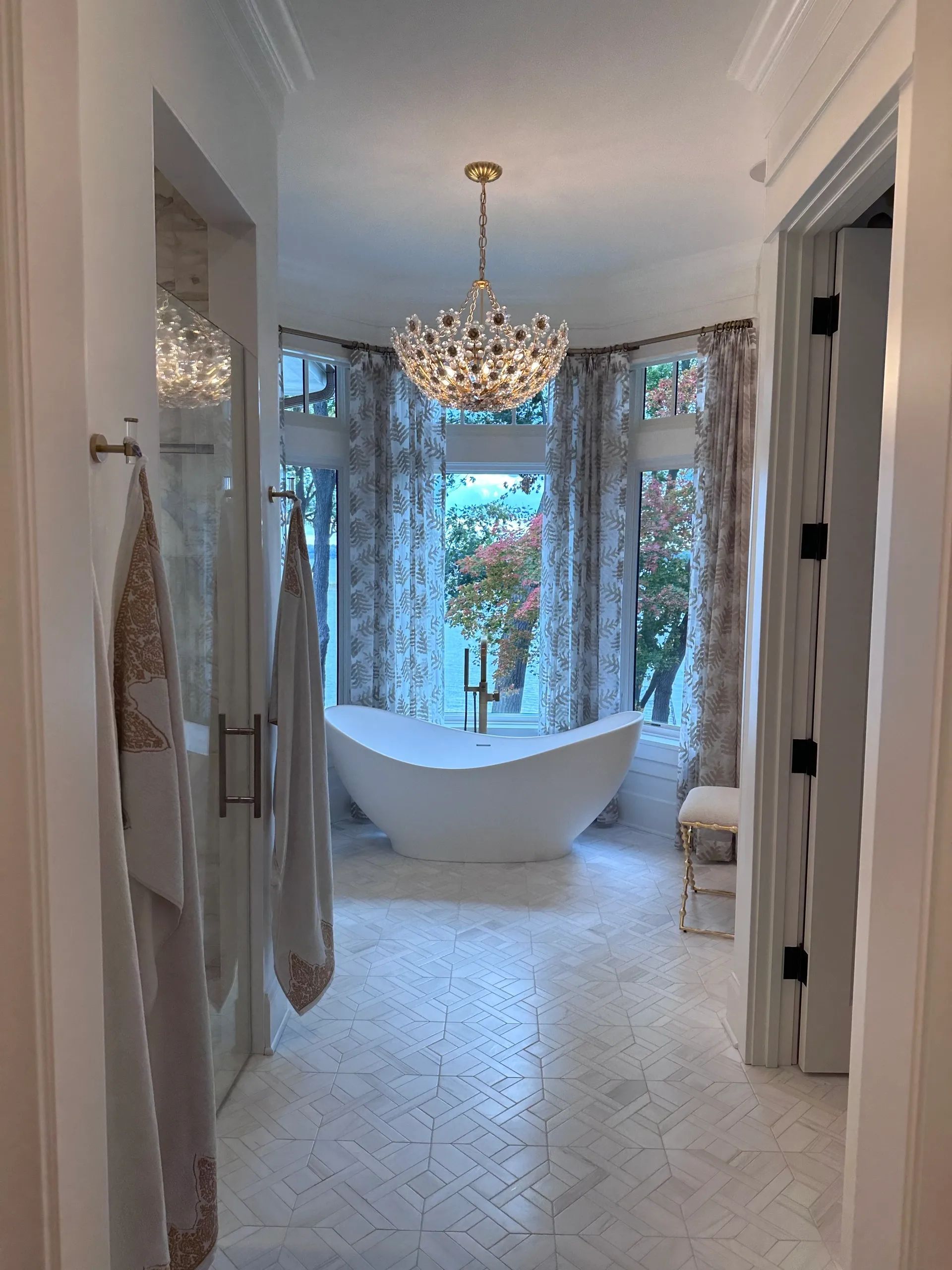 Luxurious bathroom with freestanding tub, chandelier, windows with floral curtains.