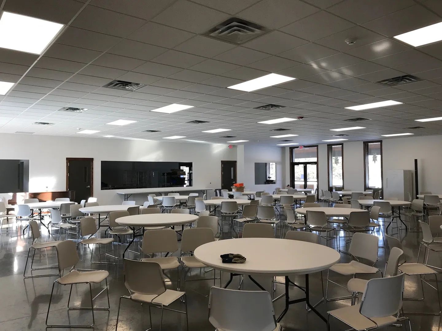 Large, empty room with round tables and chairs, possibly a cafeteria or event space. Bright lighting and gray flooring.