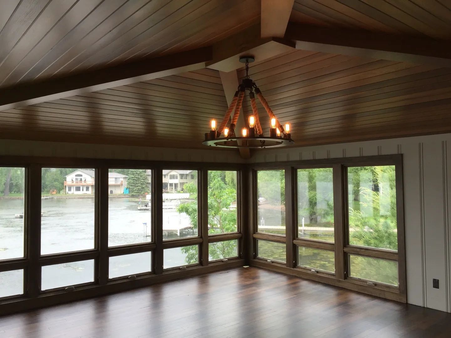 Room with large windows overlooking a lake. Wooden ceiling, chandelier, and dark wood floors.