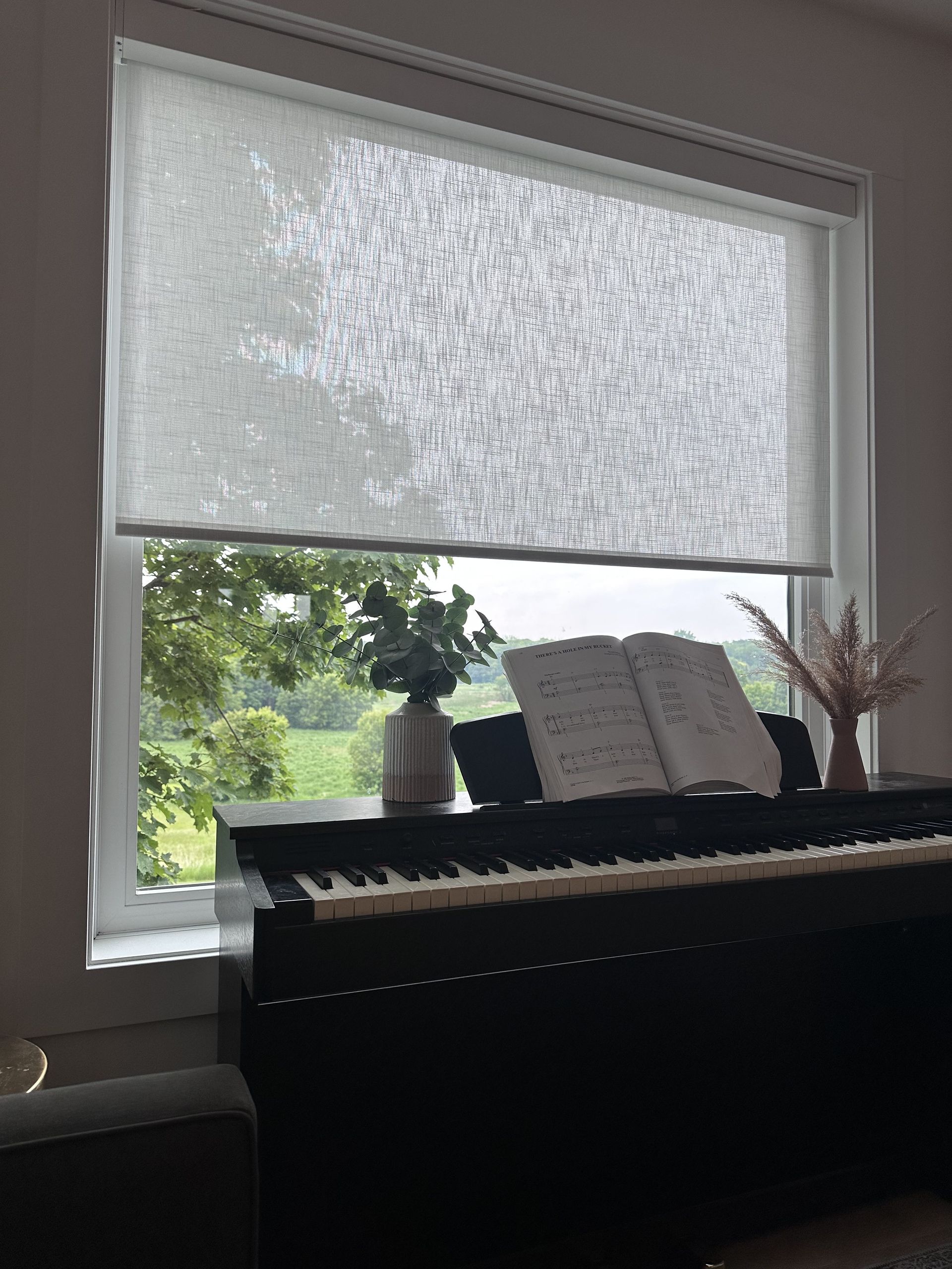 Piano by a window with a partly drawn light gray roller shade, overlooking a green outdoor view.