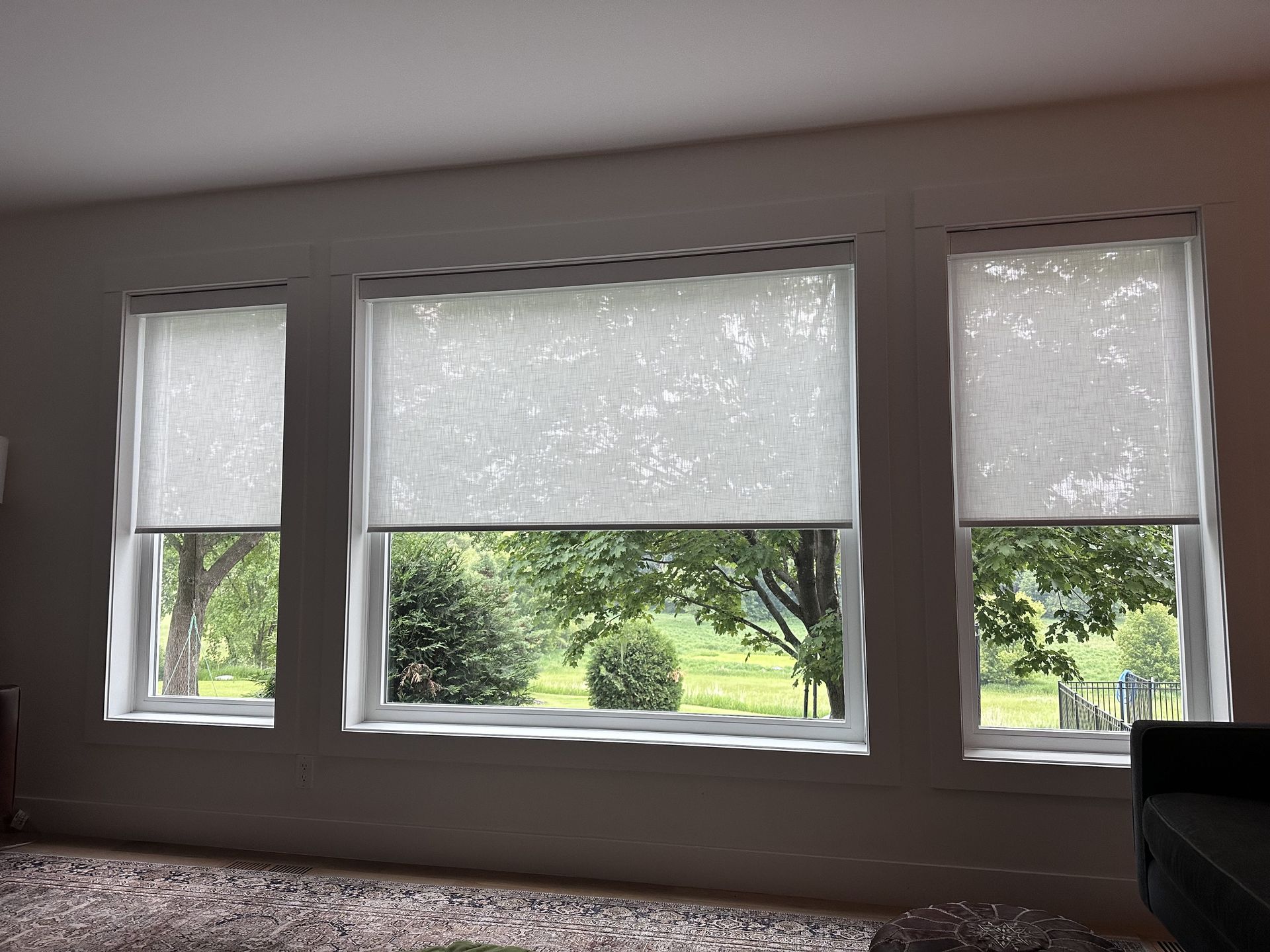 Three windows with light-colored roller shades. Outside are trees and greenery.
