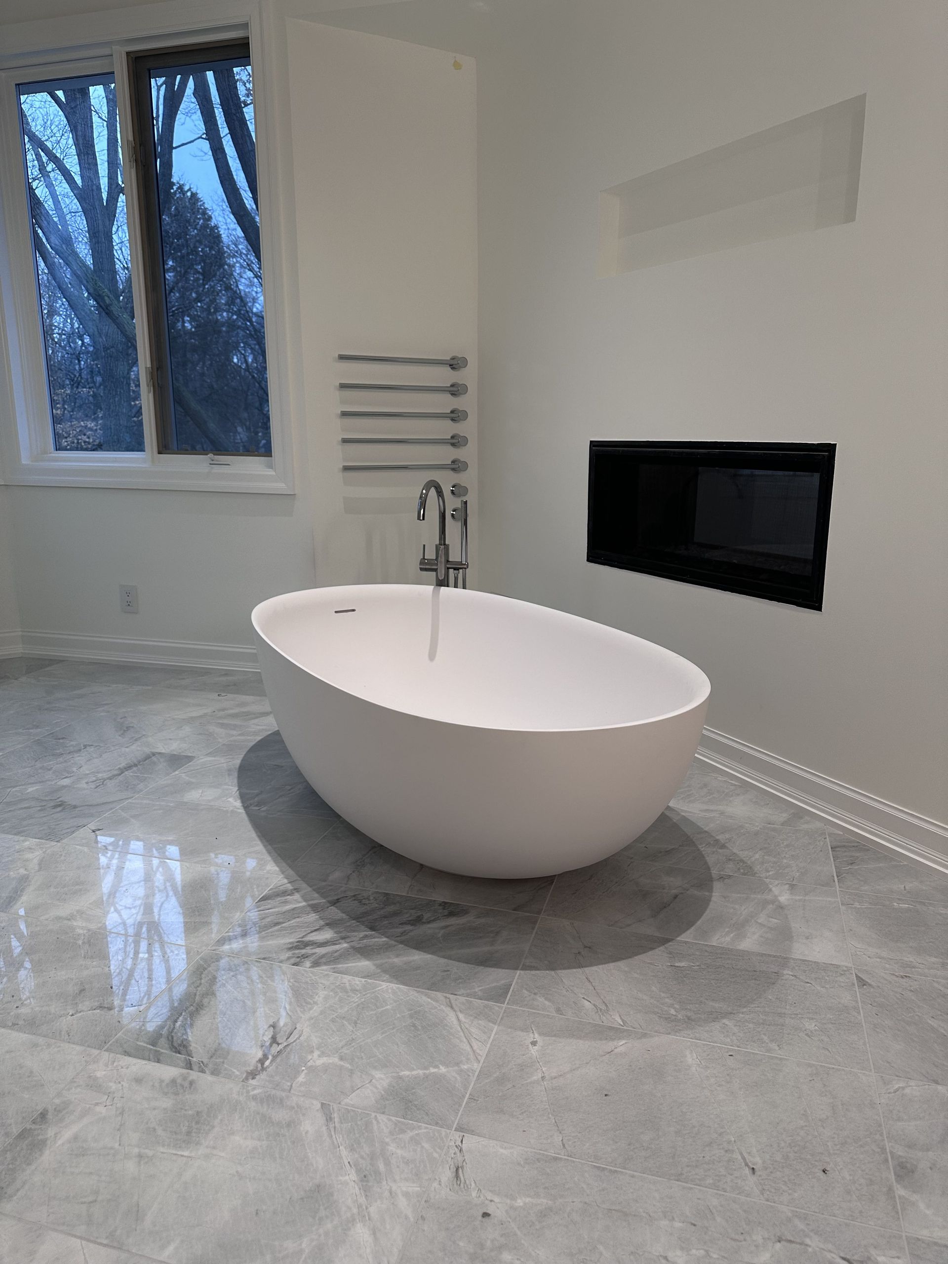 Modern white bathroom with an oval tub, gray tiled floor, and window.