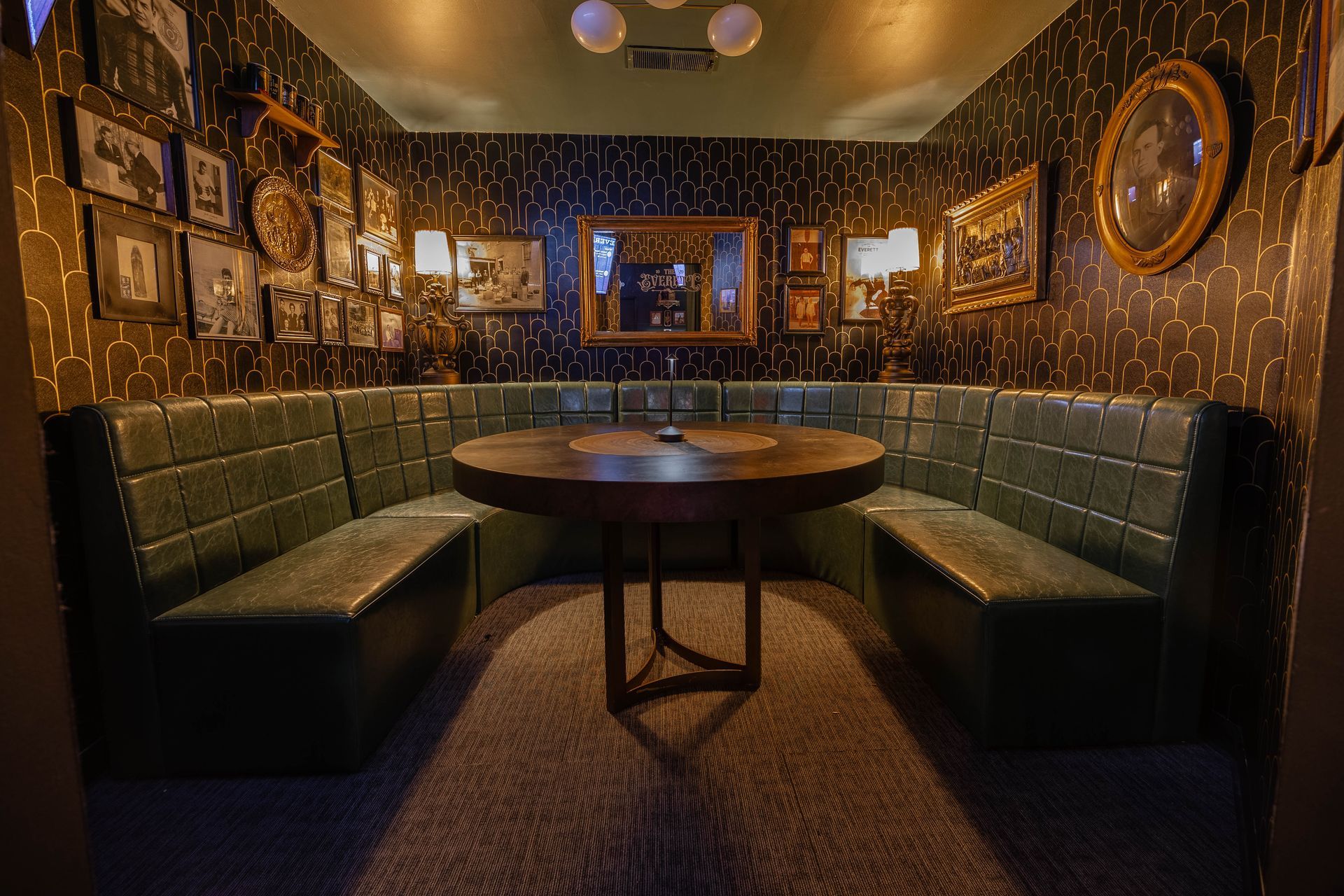 A dimly lit booth in a bar with green leather seating and a dark wood table. Artwork and a mirror decorate the walls.