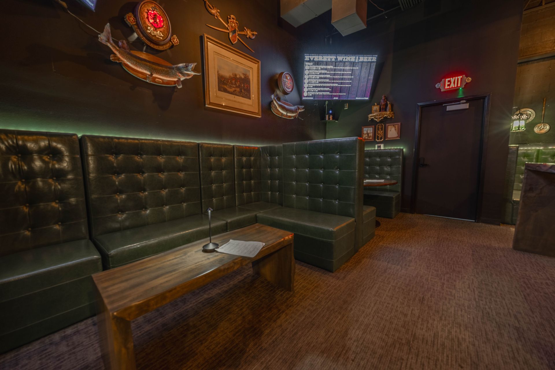 A dimly lit lounge area with a dark green tufted leather booth, wooden coffee table, and decorative wall art.
