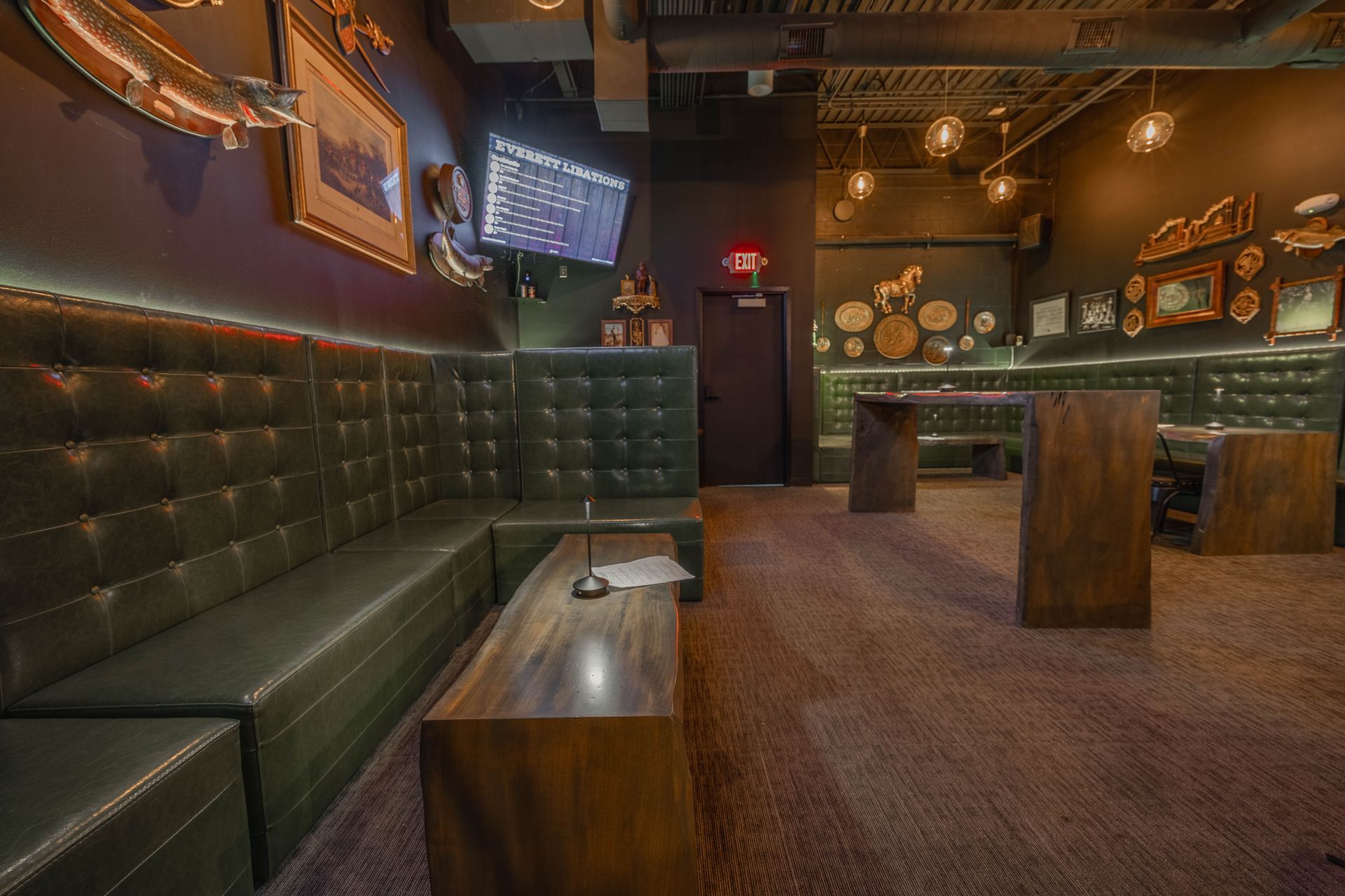 Interior of a dimly lit bar with dark green booth seating, wooden tables, and decorative wall art. Brown carpet and exposed ceiling.