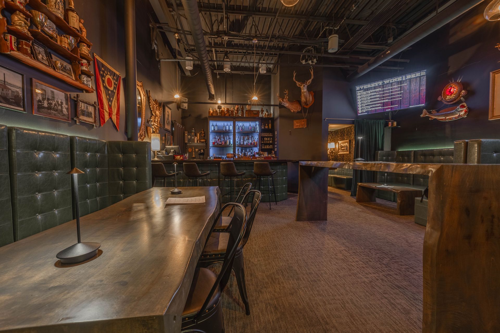 Interior view of a dimly lit bar with a long table, booths, and a bar area. Dark colors with a rustic aesthetic.