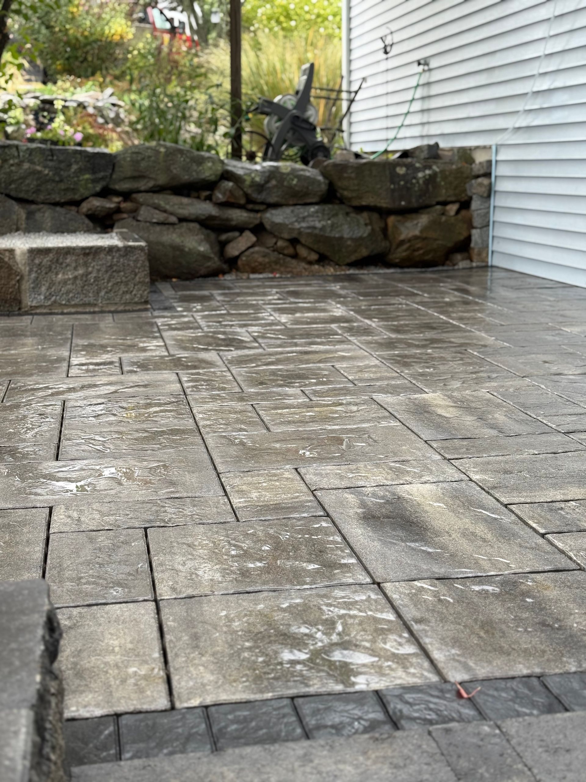 Stone patio with wet pavers beside a white house and stacked rock wall