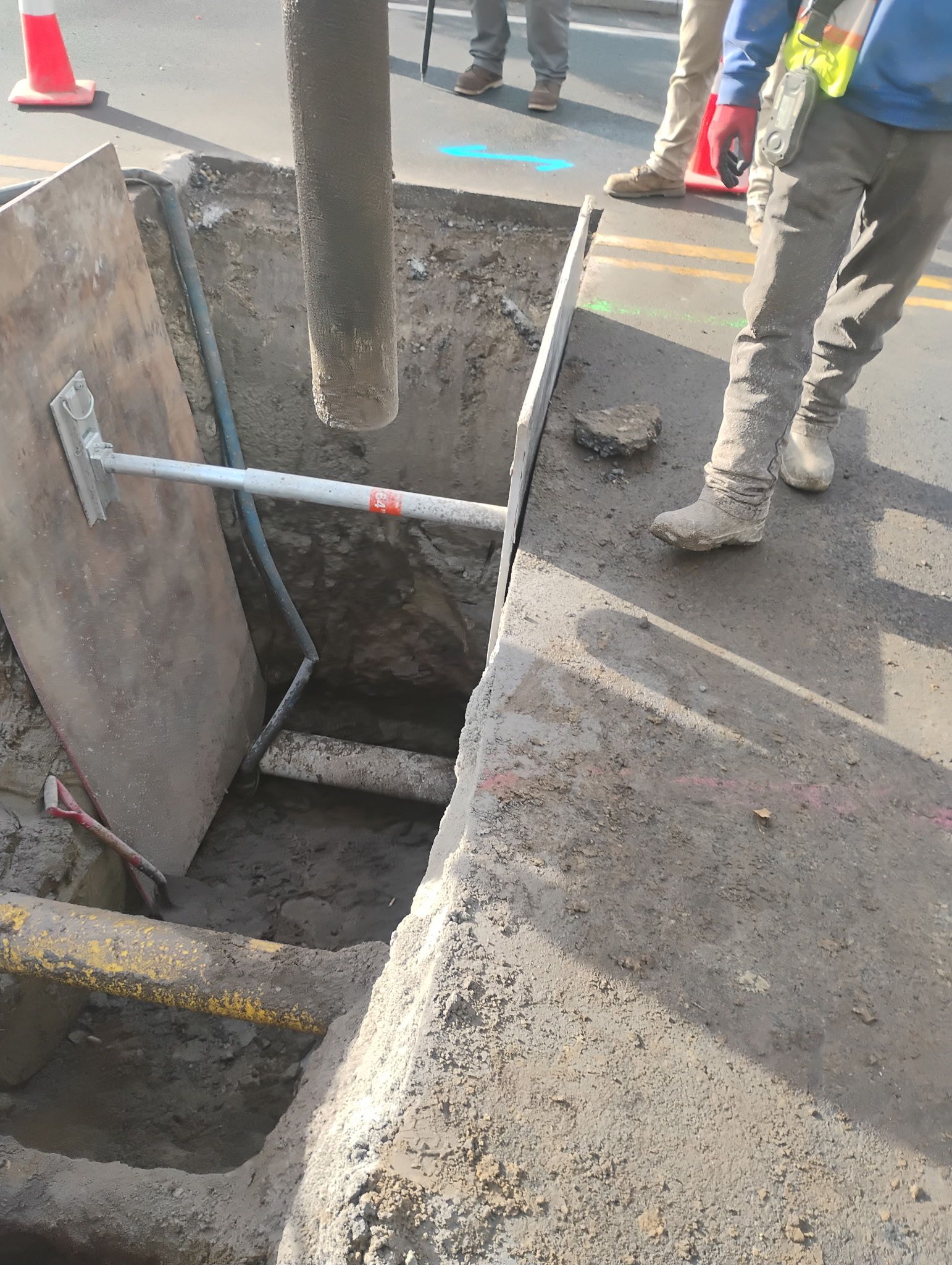 Roadwork trench with exposed pipe, metal brace, and workers’ legs beside a wet street.