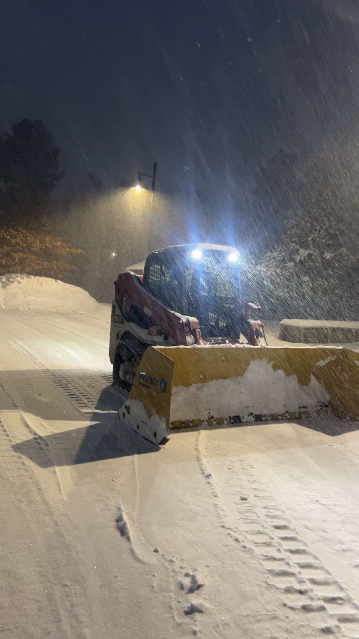 Snowplow clearing snow at night during heavy snowfall, with bright headlights and a large blade pushing snow.