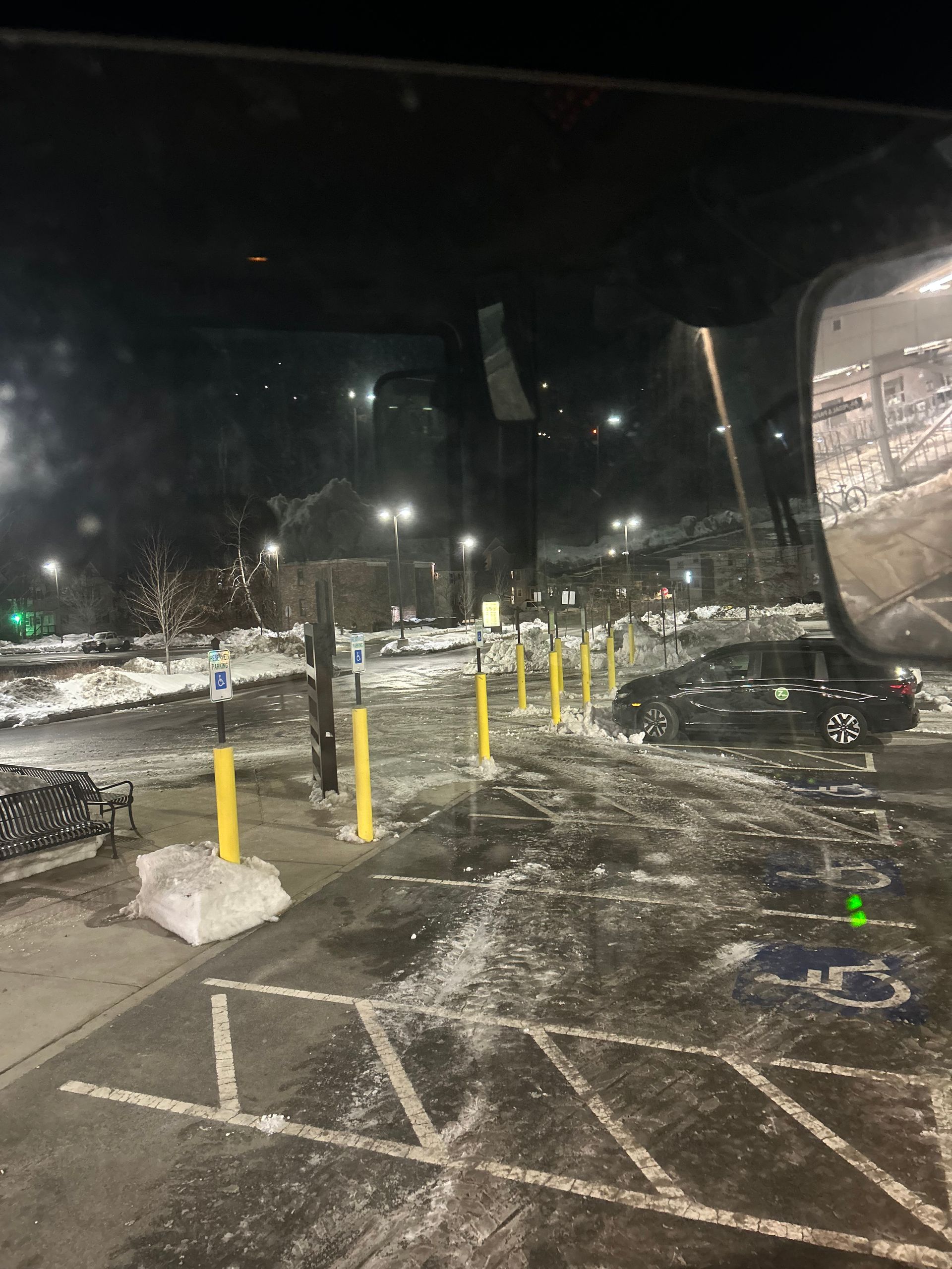 Snowy parking lot at night with yellow bollards, parked cars, and a wet, slushy ground