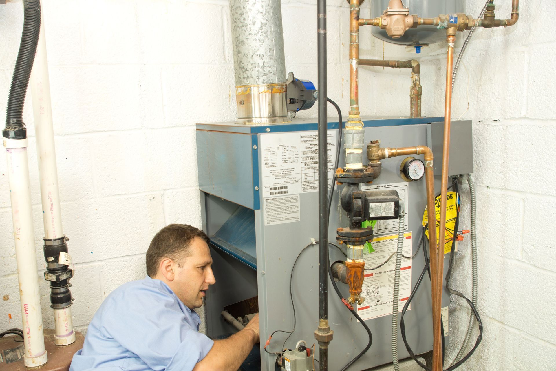 Technician inspecting a blue furnace in a utility room with exposed pipes and equipment