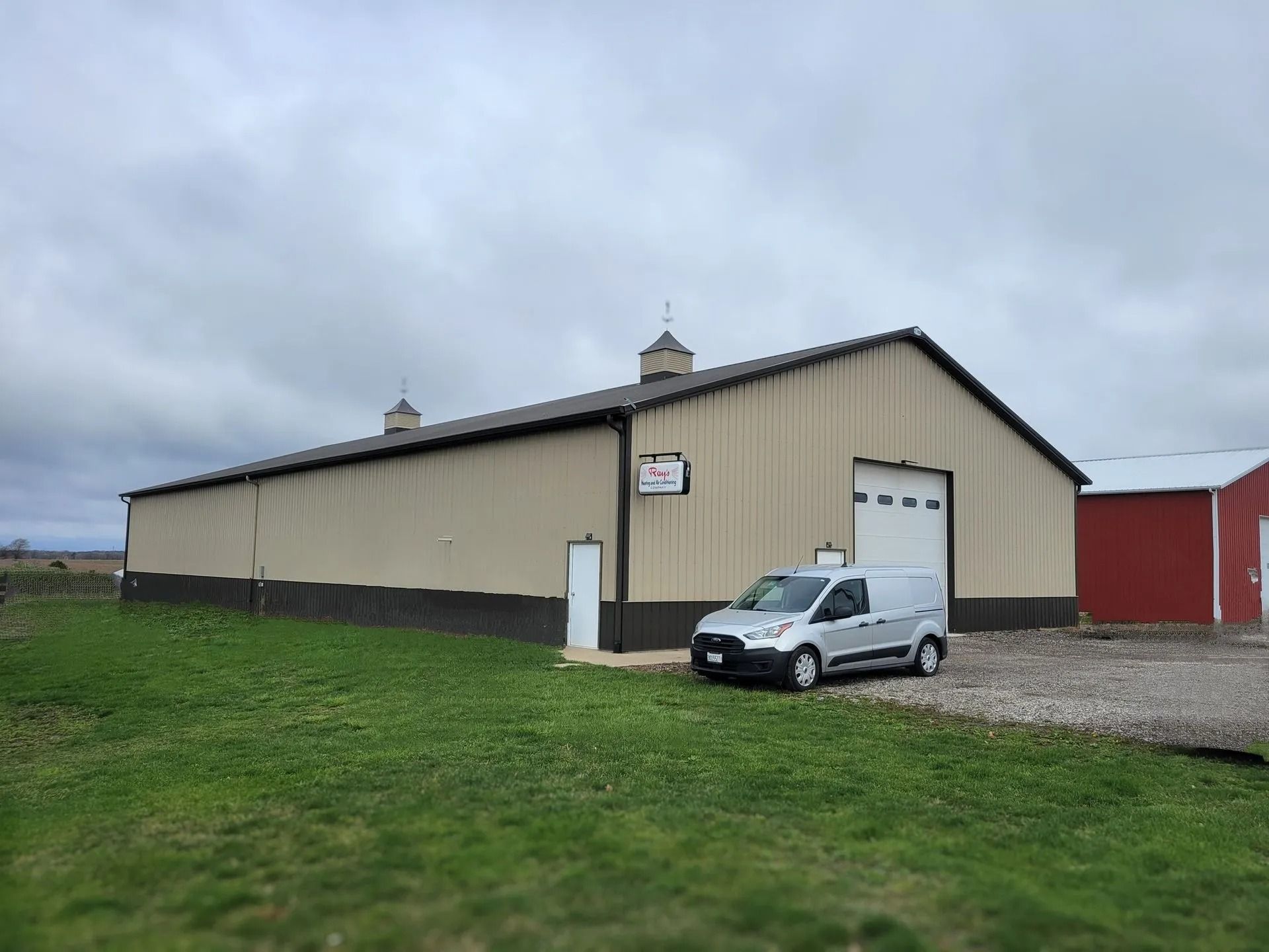 Tan metal barn with a white door and a small silver car parked outside on gravel.