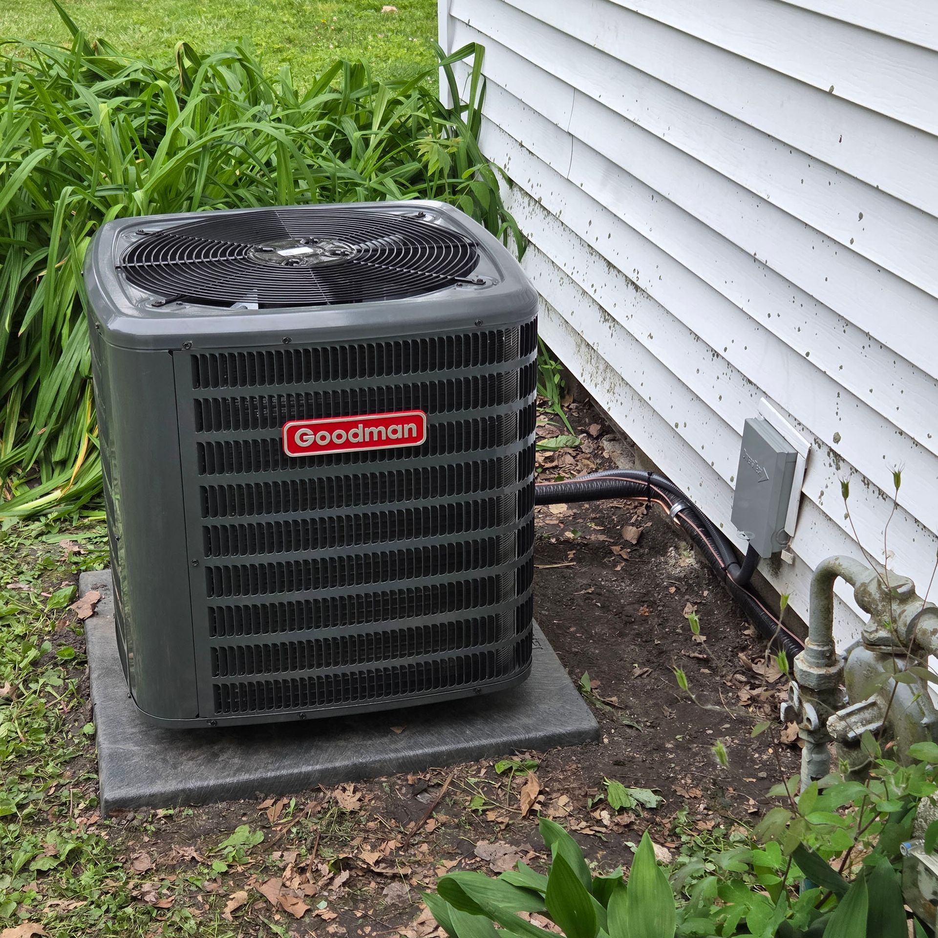 Goodman outdoor air conditioner unit beside a house with white siding and overgrown grass