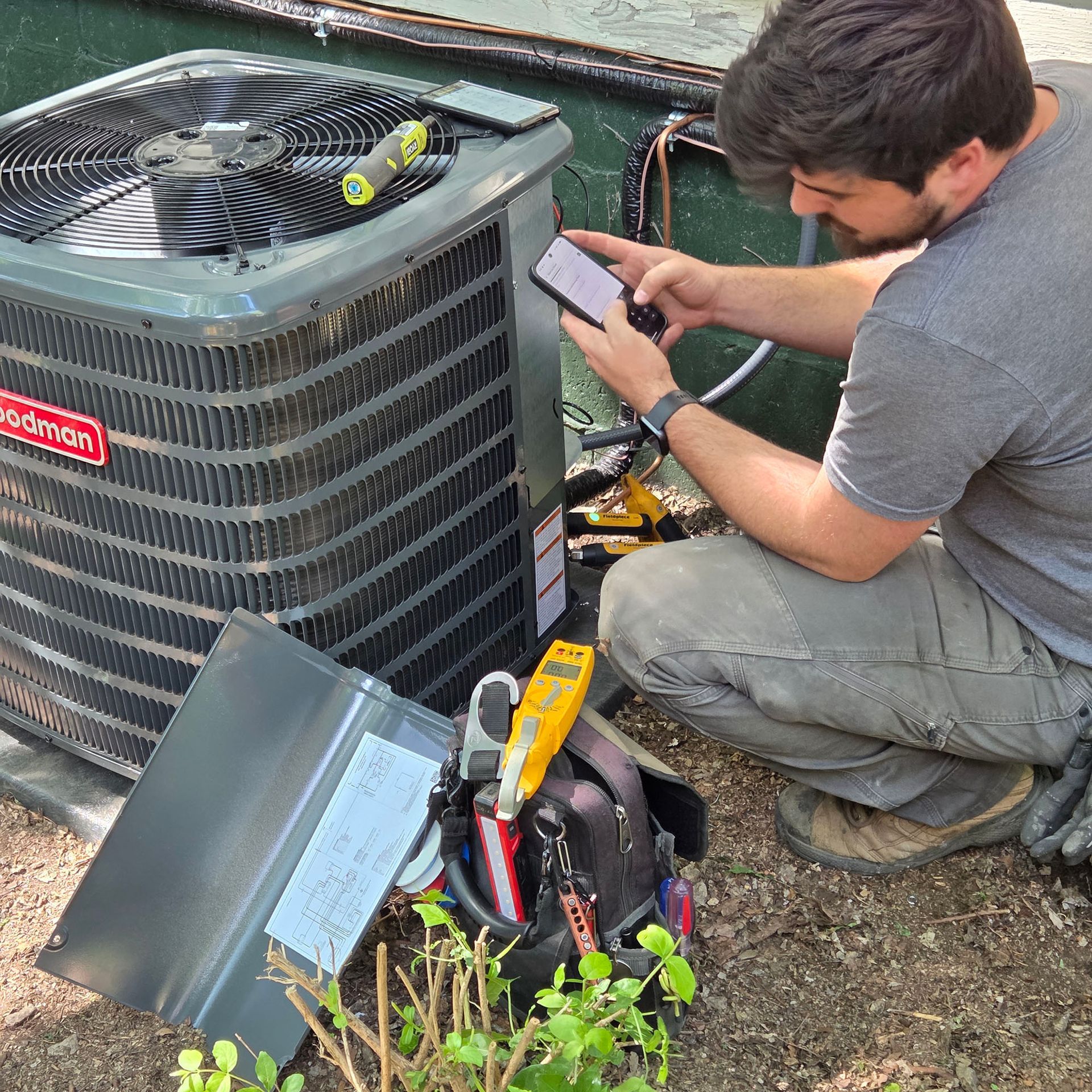 Technician inspecting a damaged outdoor air conditioner with a phone and open tool bag.