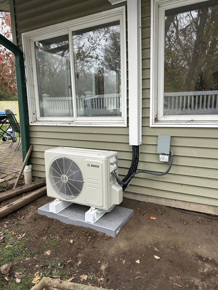 Outdoor air conditioner condenser beside a house window on a concrete pad, with white siding and trees behind.