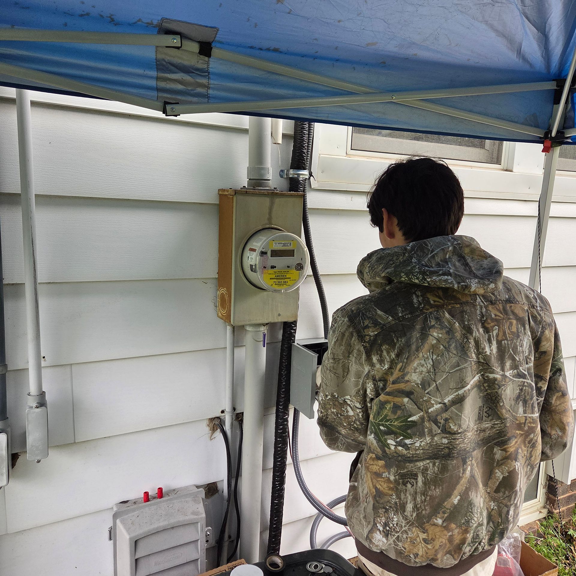 Person in camouflage jacket inspecting an electric meter under a blue canopy outdoors.
