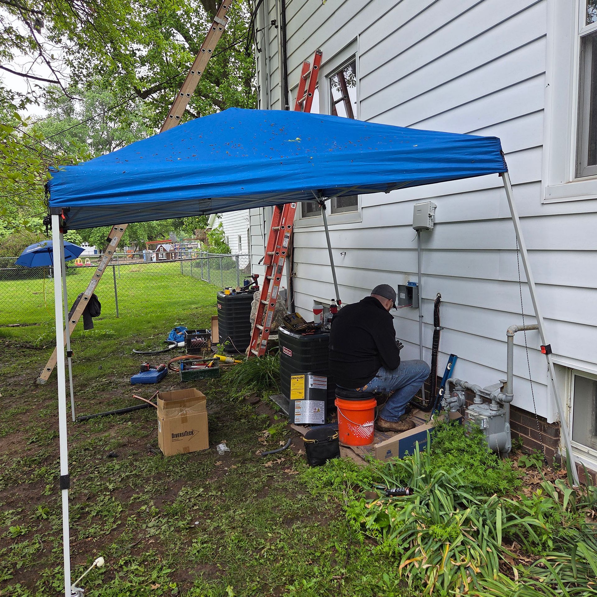 Blue canopy tent beside a white house, with a person working on the ground and tools scattered nearby.