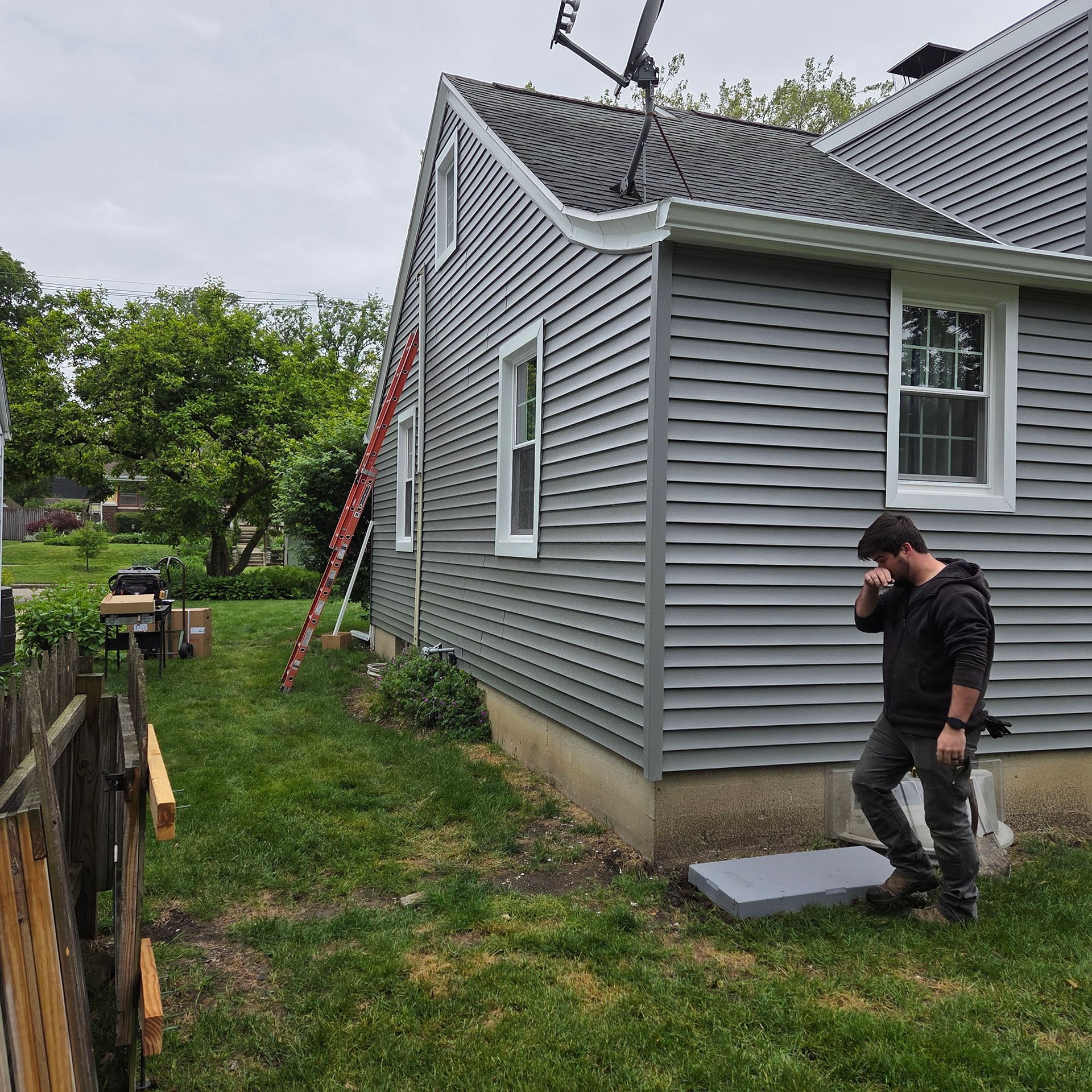 Man standing by gray house with a ladder, inspecting the exterior near the foundation.