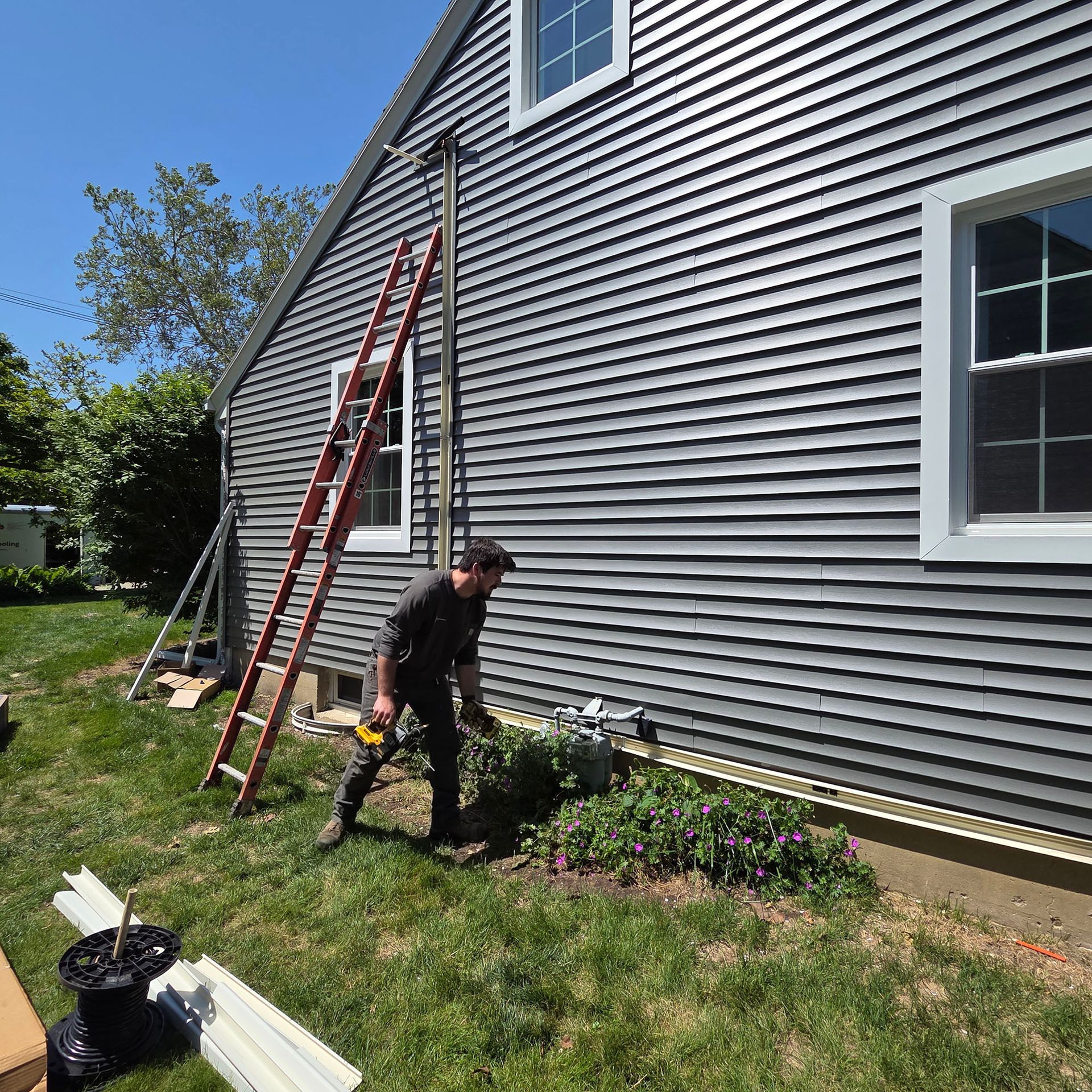 Worker trimming plants beside a gray house, with a red ladder leaning against the wall.