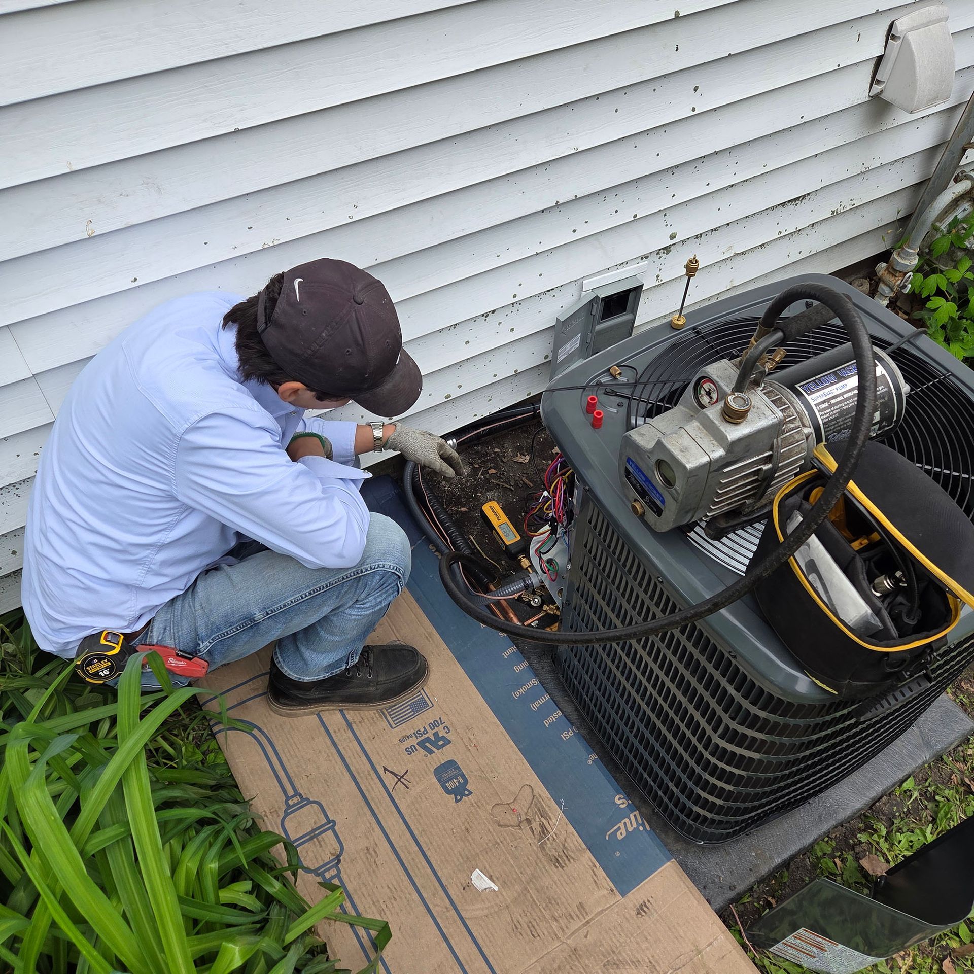 Technician kneels beside an outdoor HVAC unit, inspecting wiring and components next to a house.