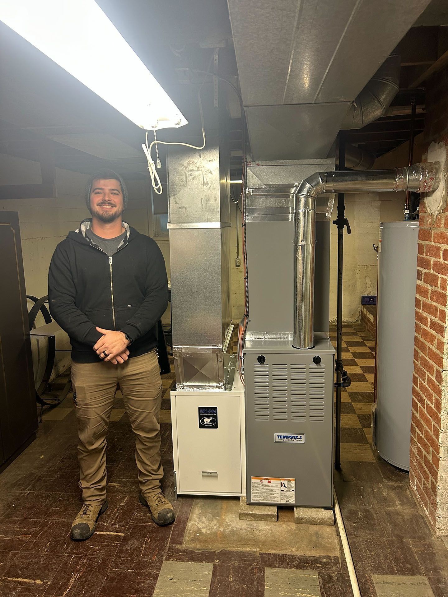 Man standing in a utility basement beside a furnace, boiler, and exposed pipes under bright fluorescent lights
