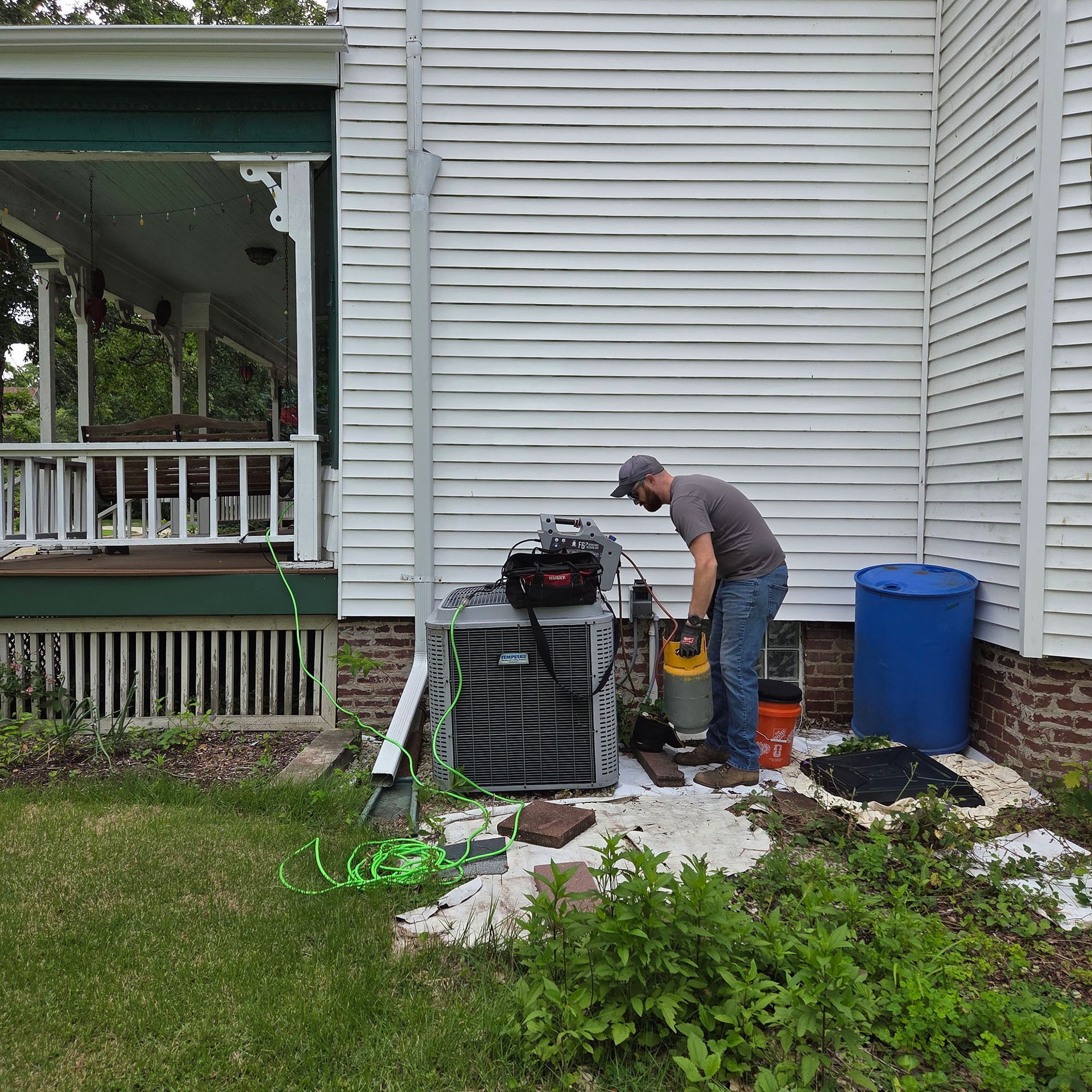 Person working beside house HVAC unit and blue rain barrel in a small yard, with hoses and equipment.