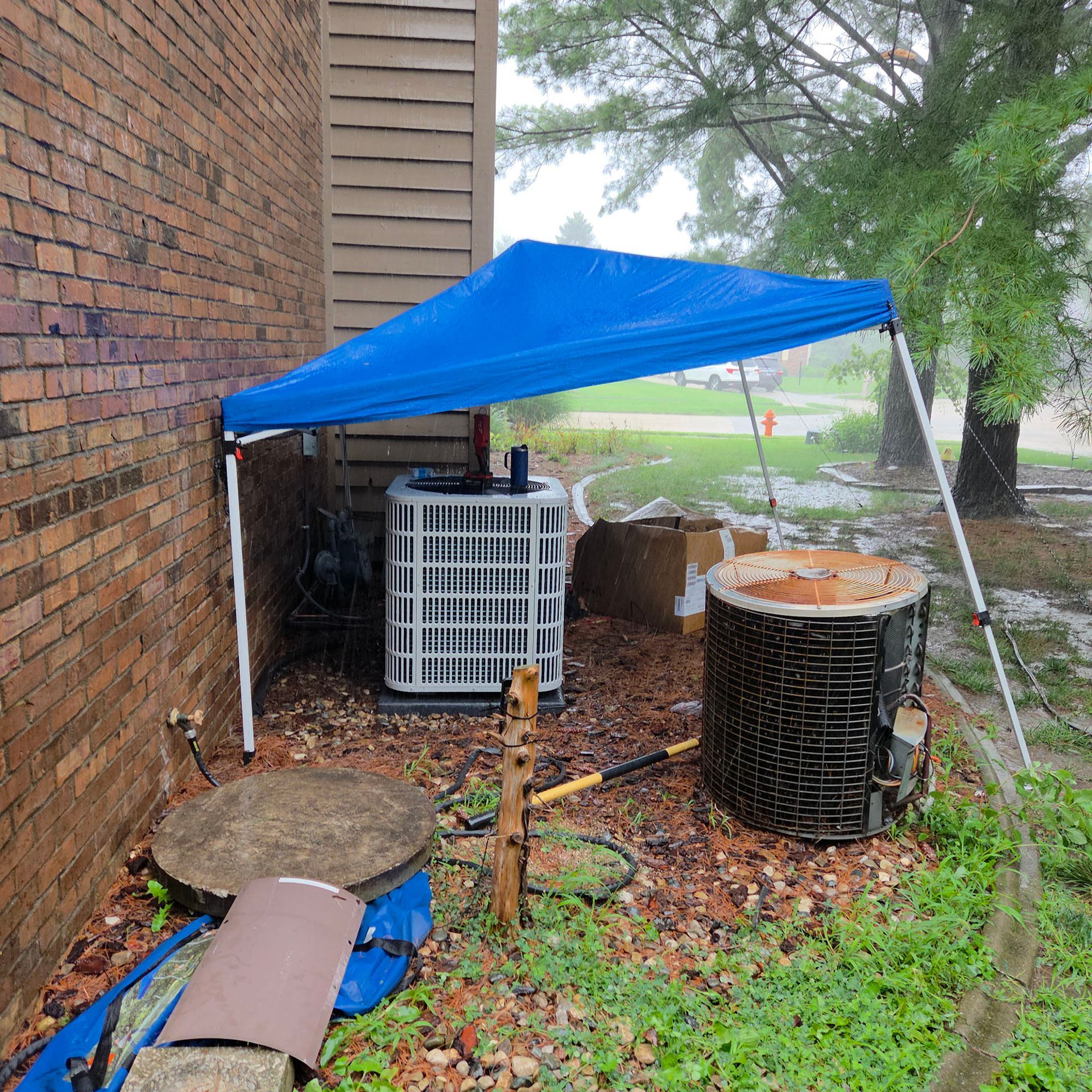 Blue canopy tent beside a brick wall shelters outdoor equipment and debris on a wet ground.