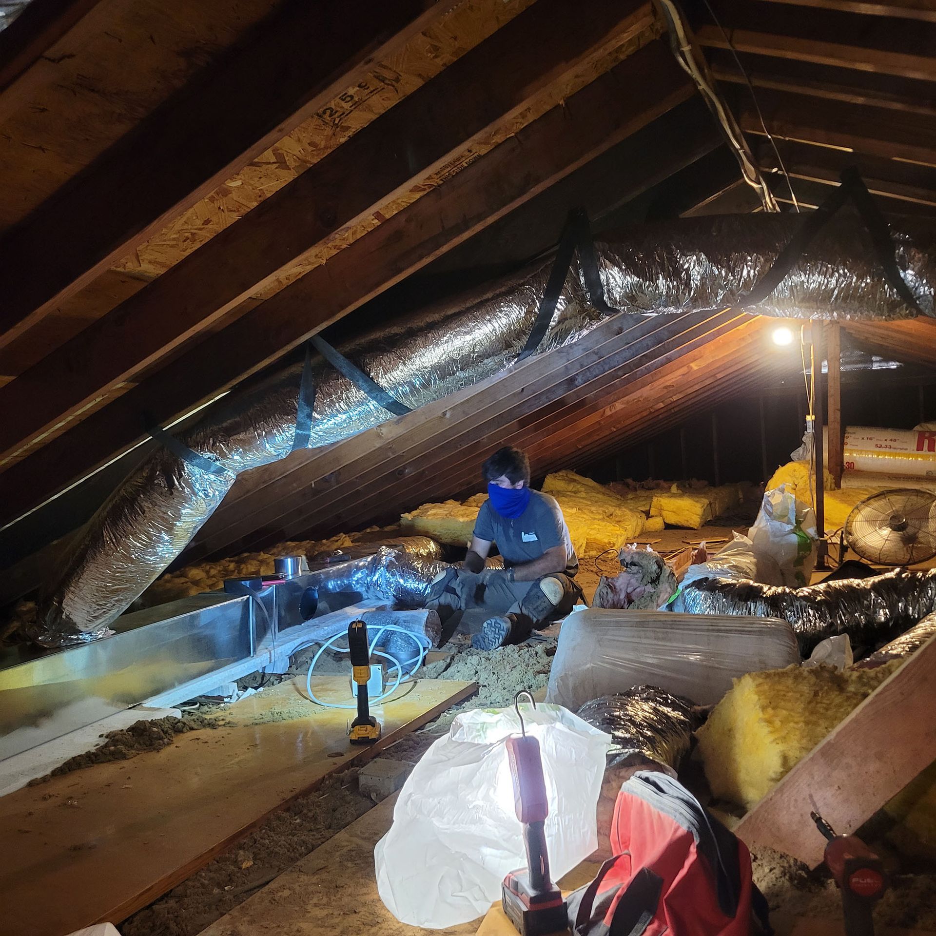 Attic crawlspace with exposed beams, plastic sheeting, and a worker installing insulation under warm light