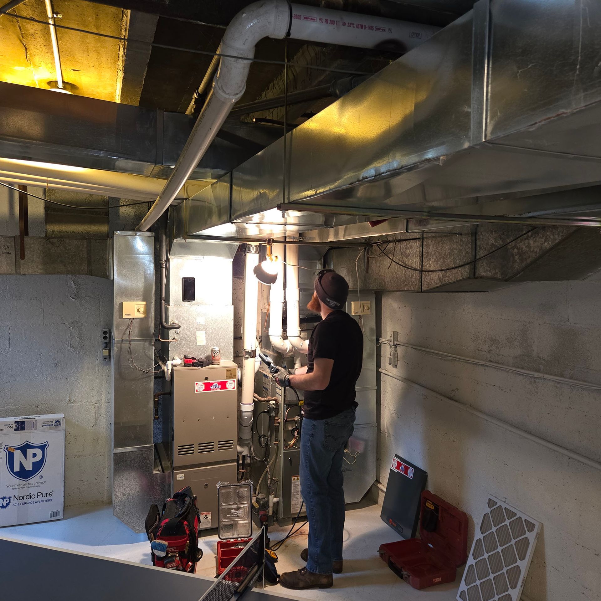 Technician inspecting HVAC equipment in a basement utility room with ductwork and exposed pipes.