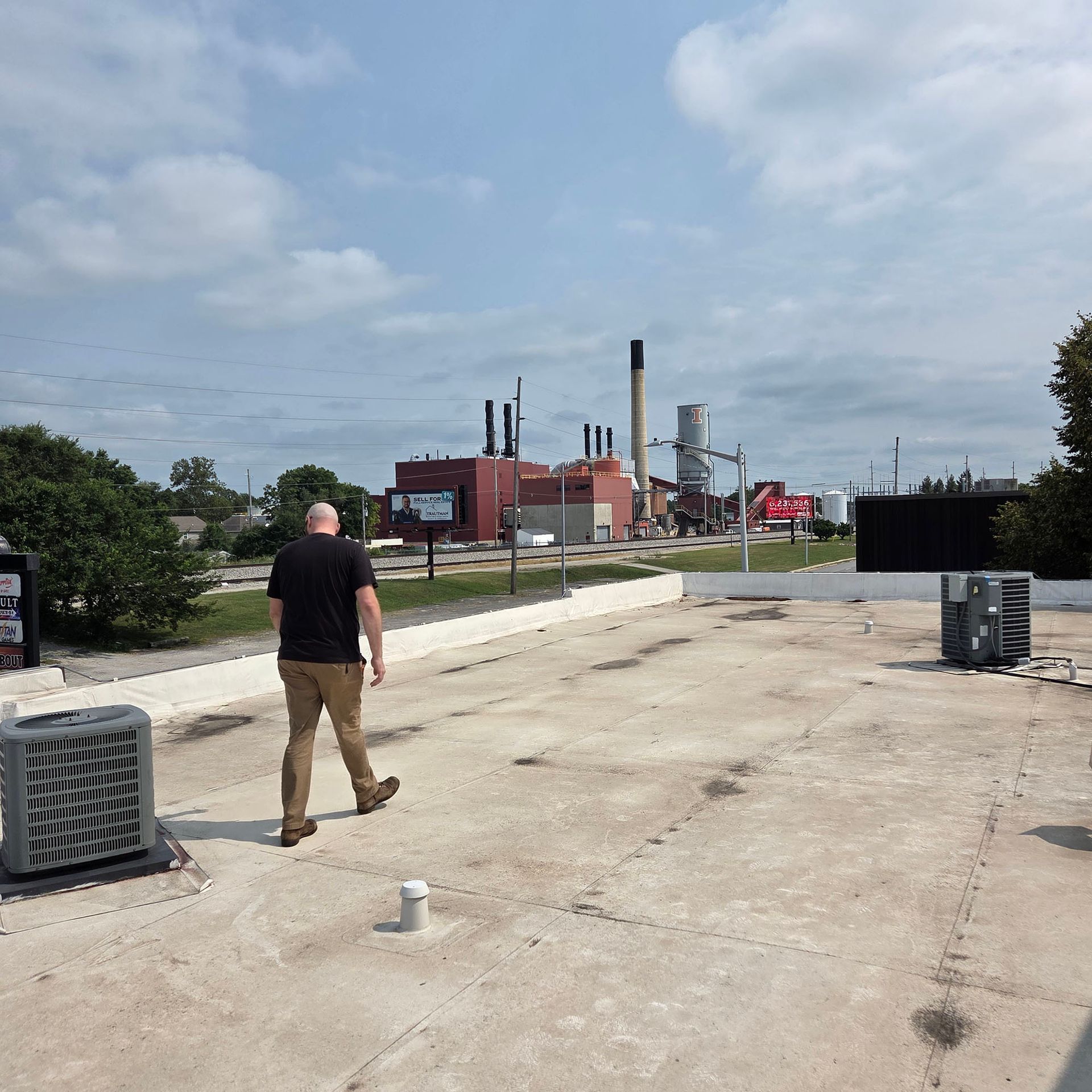 Person walking on a concrete lot near industrial buildings and smokestacks under a partly cloudy sky