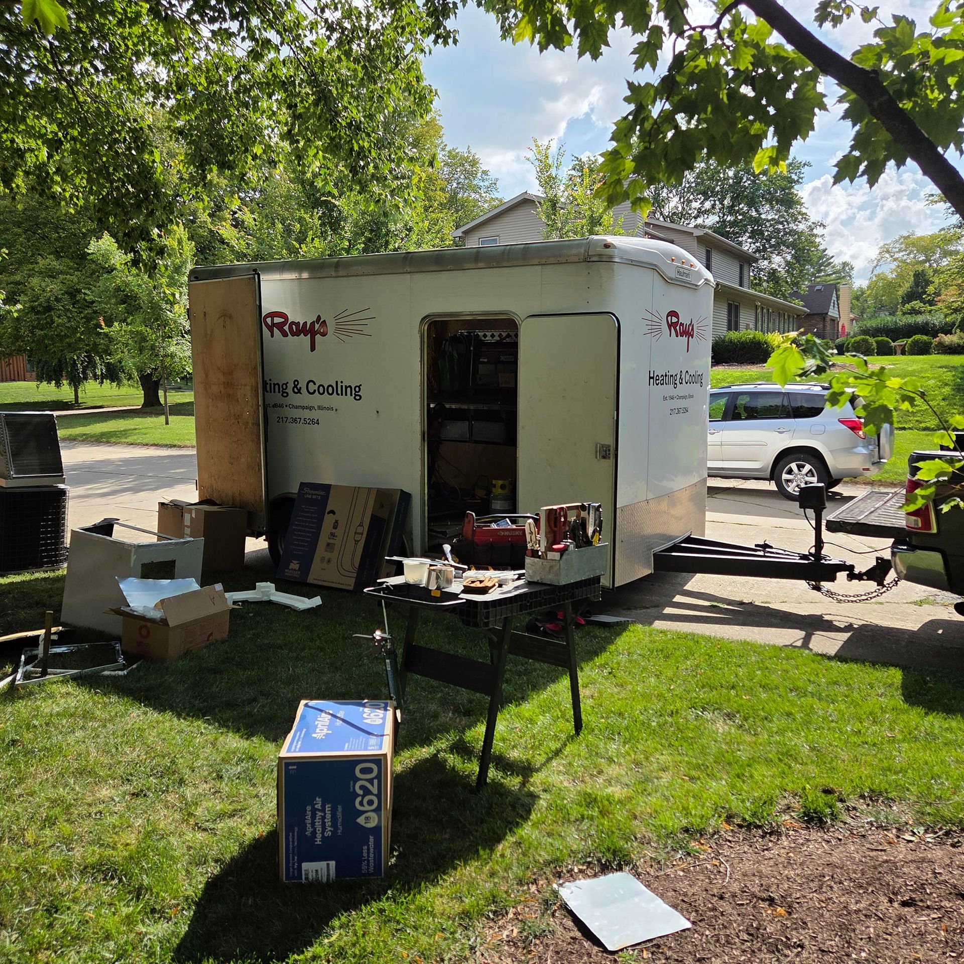 Food truck setup in a park, with tables, boxes, and equipment under trees on a sunny day