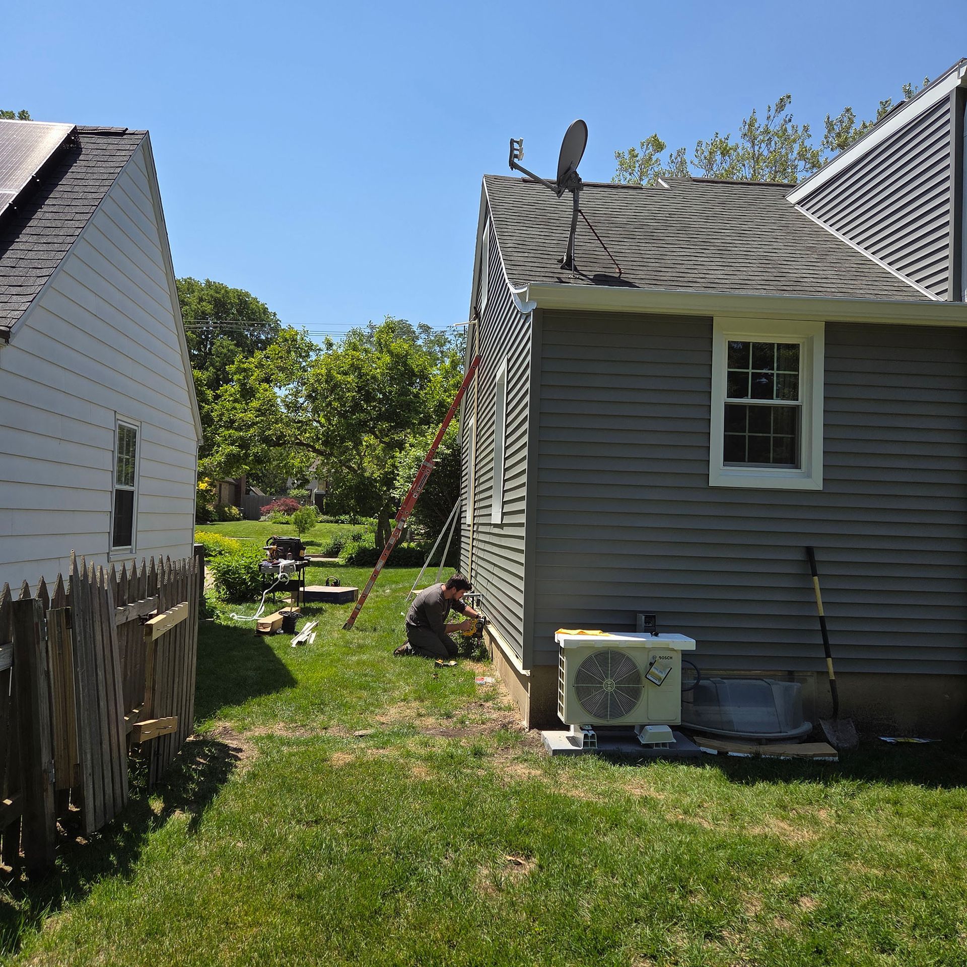 Backyard between two gray houses with a person kneeling near a ladder and lawn equipment.