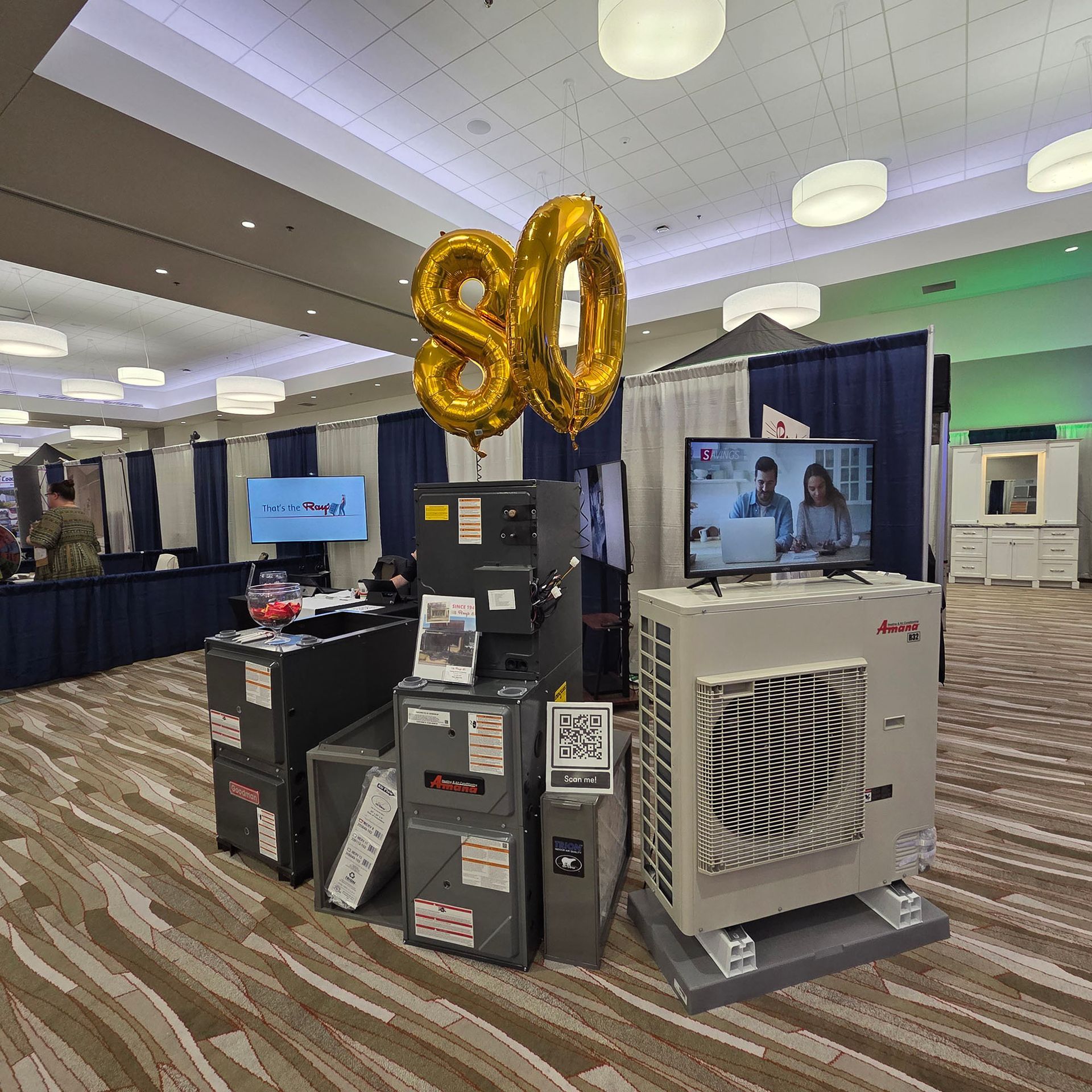 Event display with gold “80” balloons, stacked boxes, and a TV in a conference hall