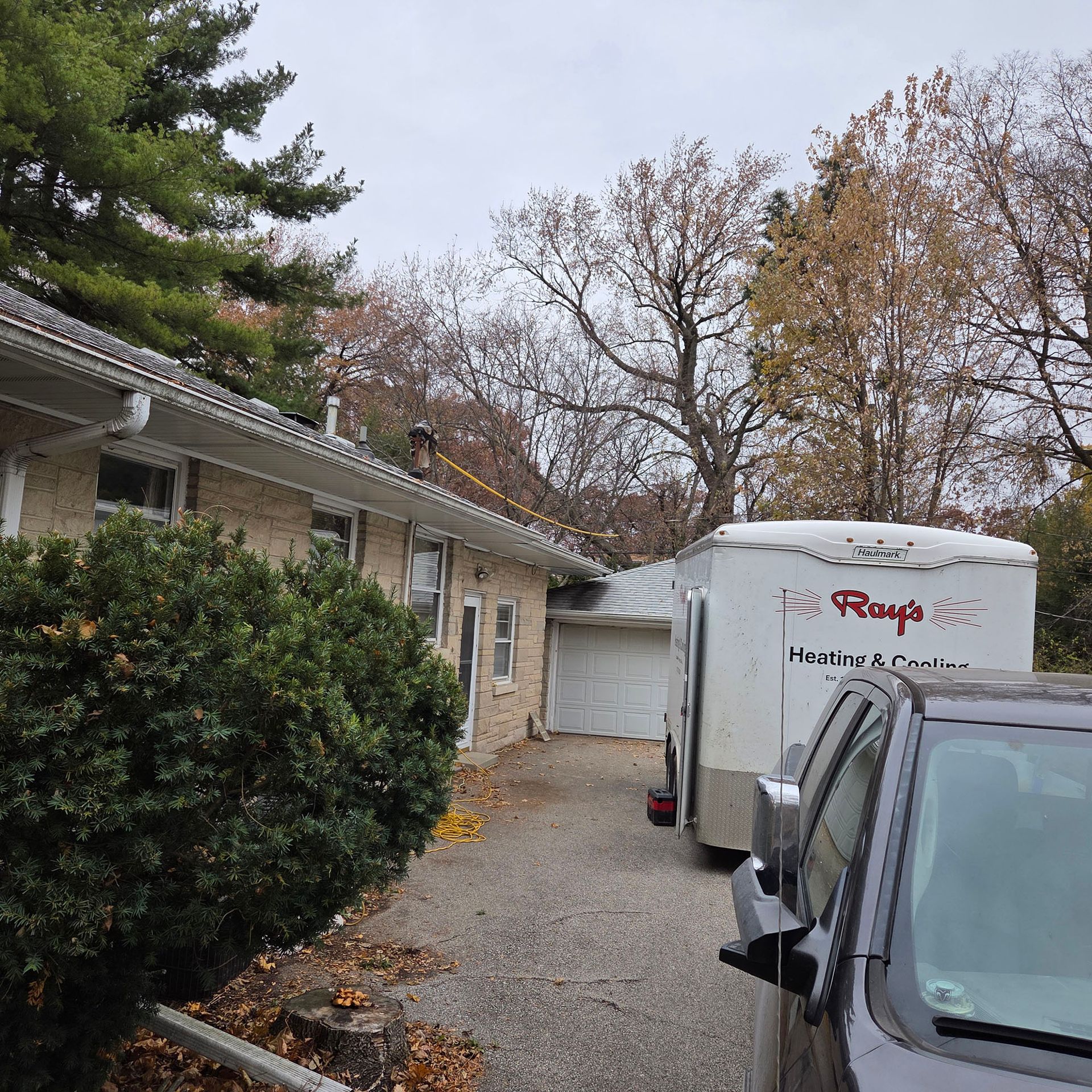 Driveway beside a house with parked vehicles, a trailer, and autumn trees under a cloudy sky