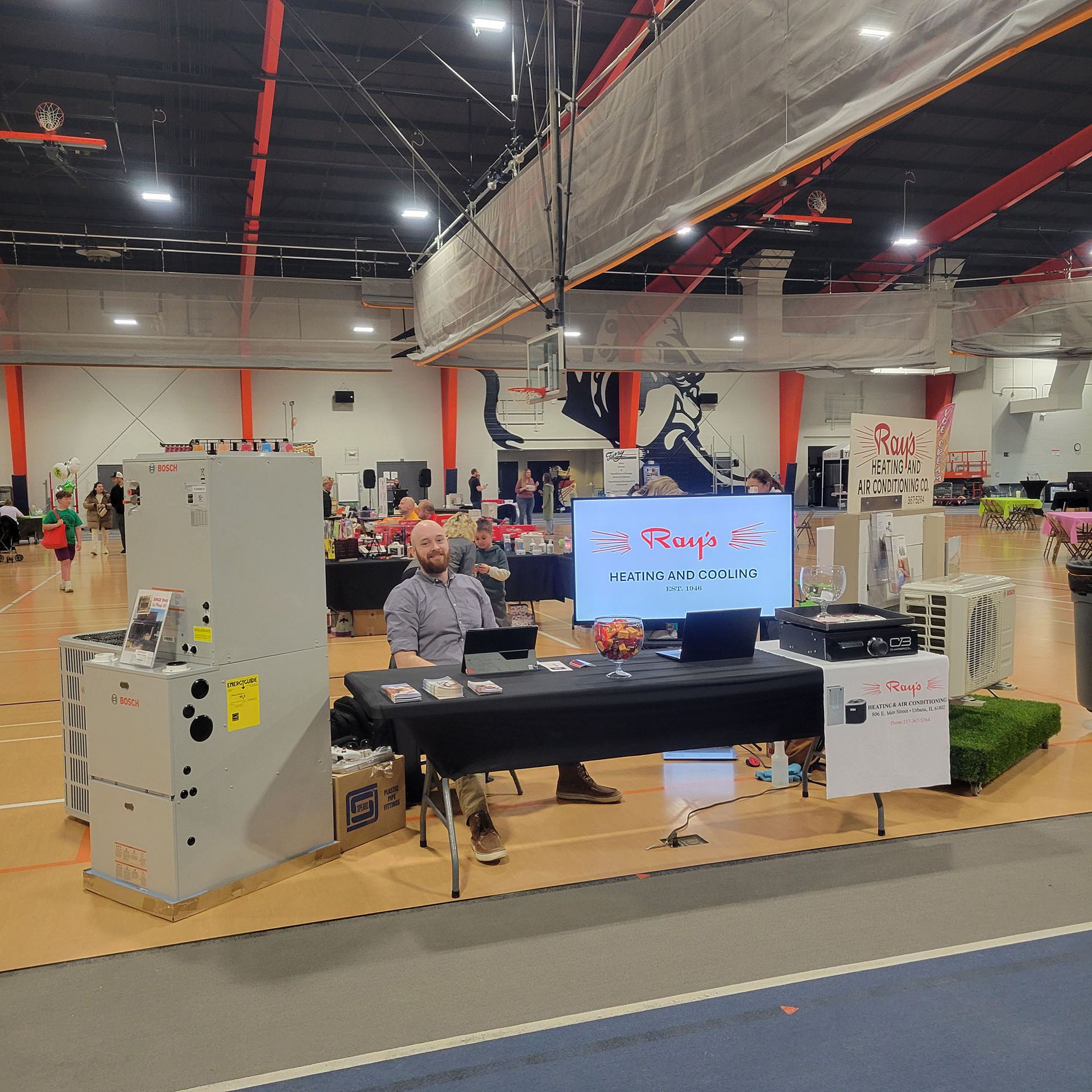 Indoor expo booth with a seated speaker, black table, and large monitor in a bright convention hall