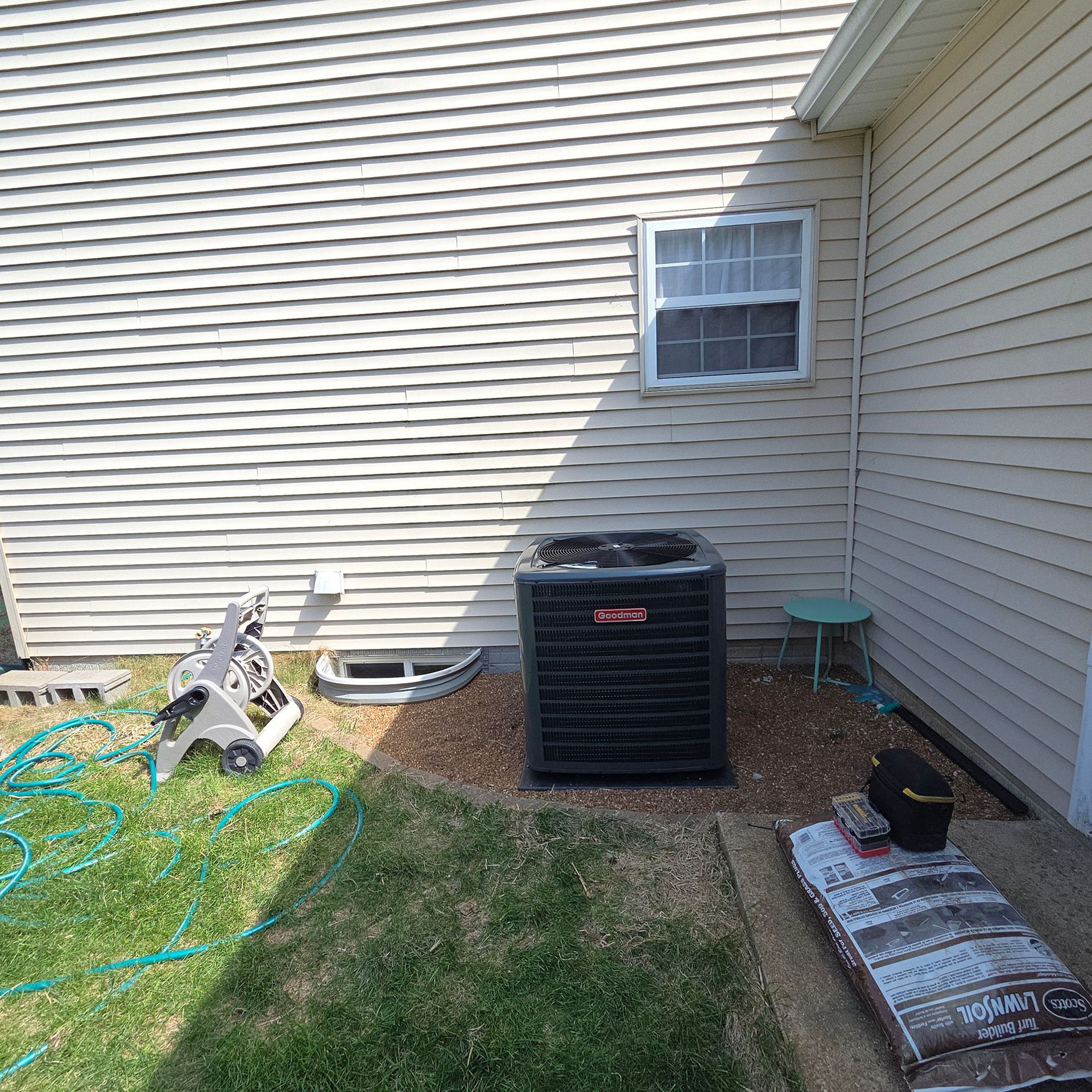 Side yard with an air conditioner unit, garden hose, and small window on a beige house.