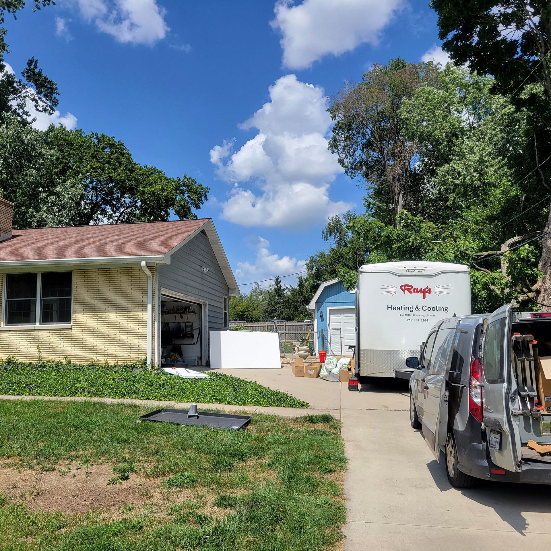 Suburban driveway beside a yellow house, with a white moving truck and parked black van under a blue sky.
